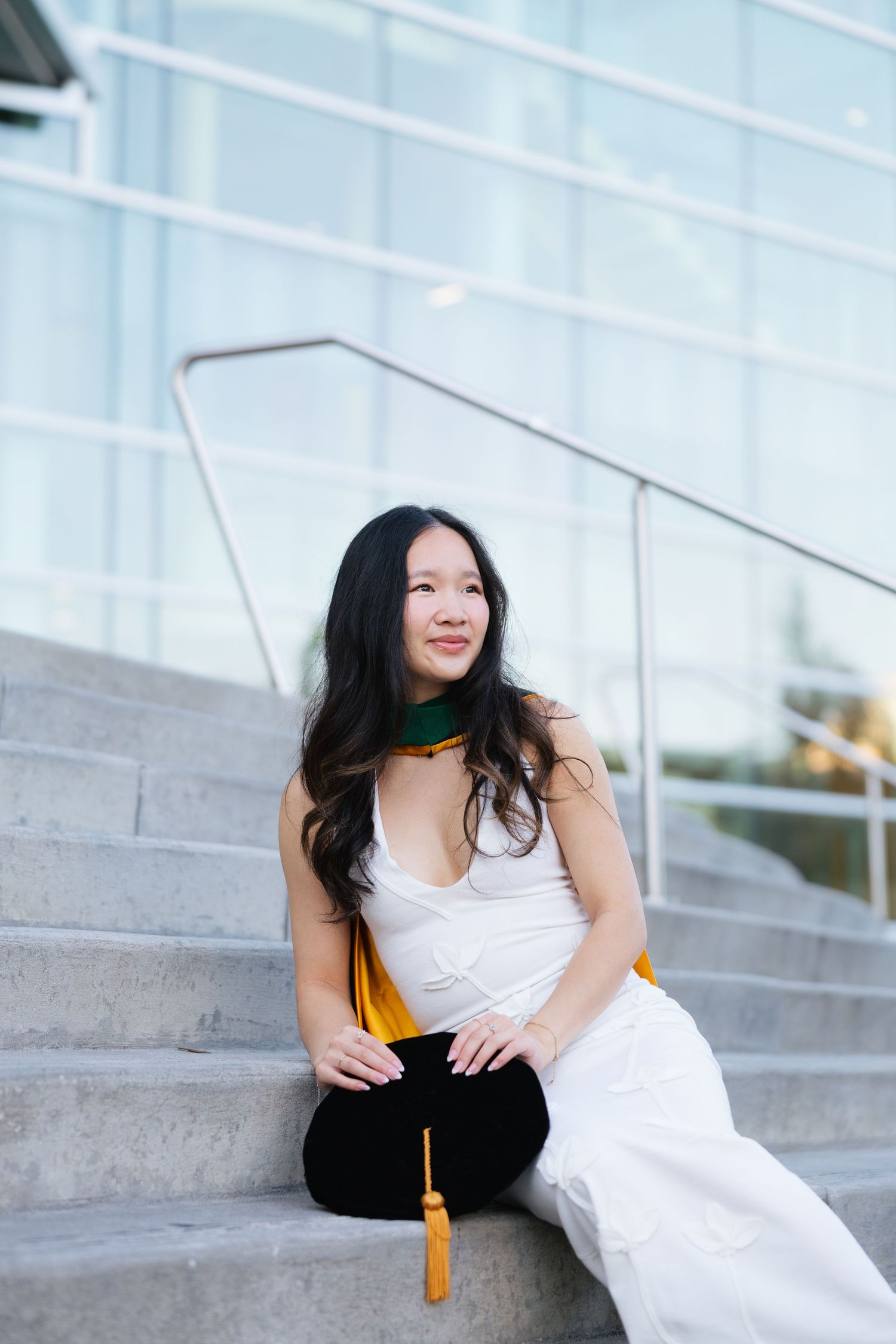 Young woman in white outfit sitting on steps holding graduation cap with gold tassel and green hood.