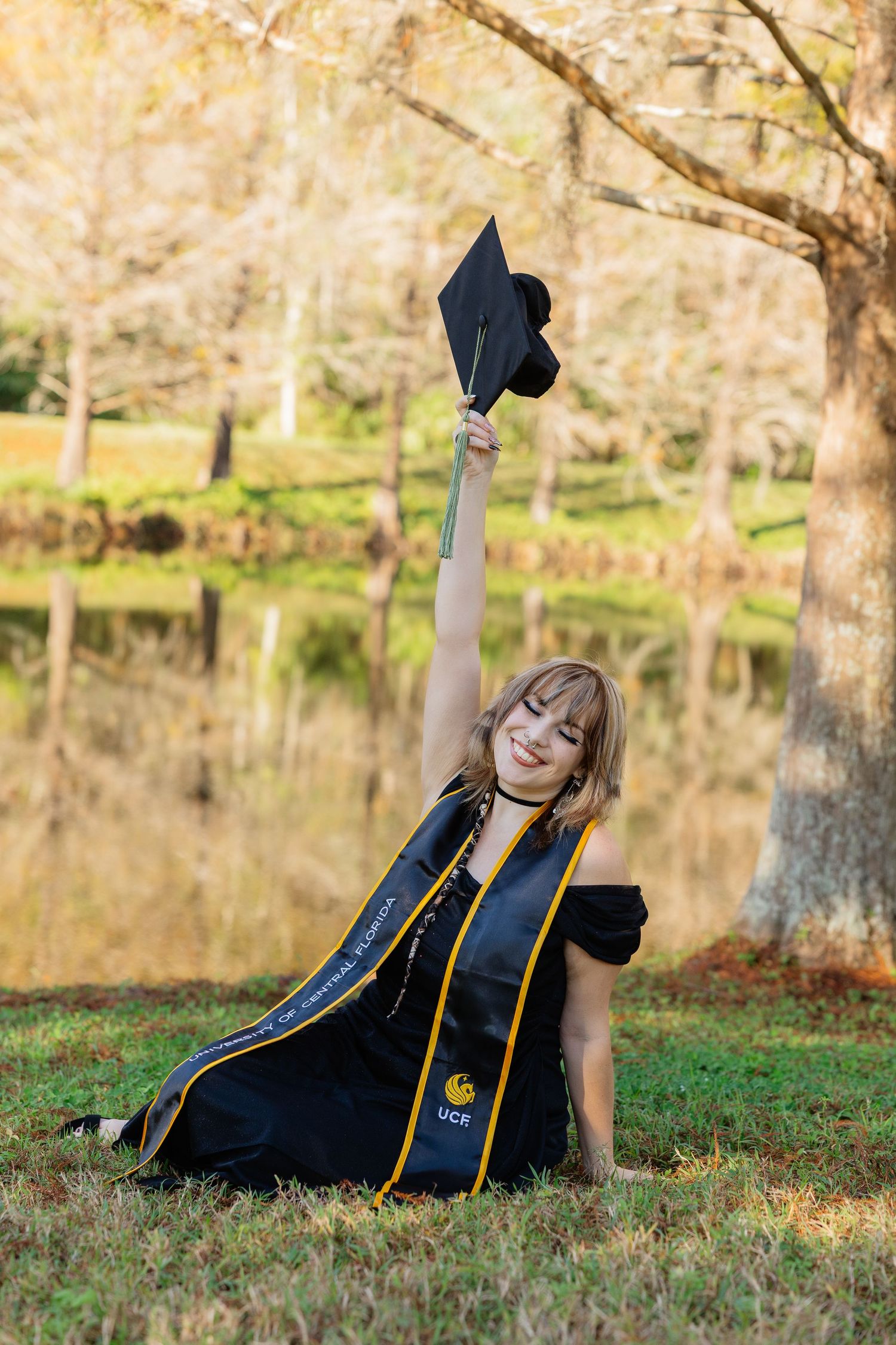 Graduate in black academic regalia poses on grass near a reflective pond during golden hour.