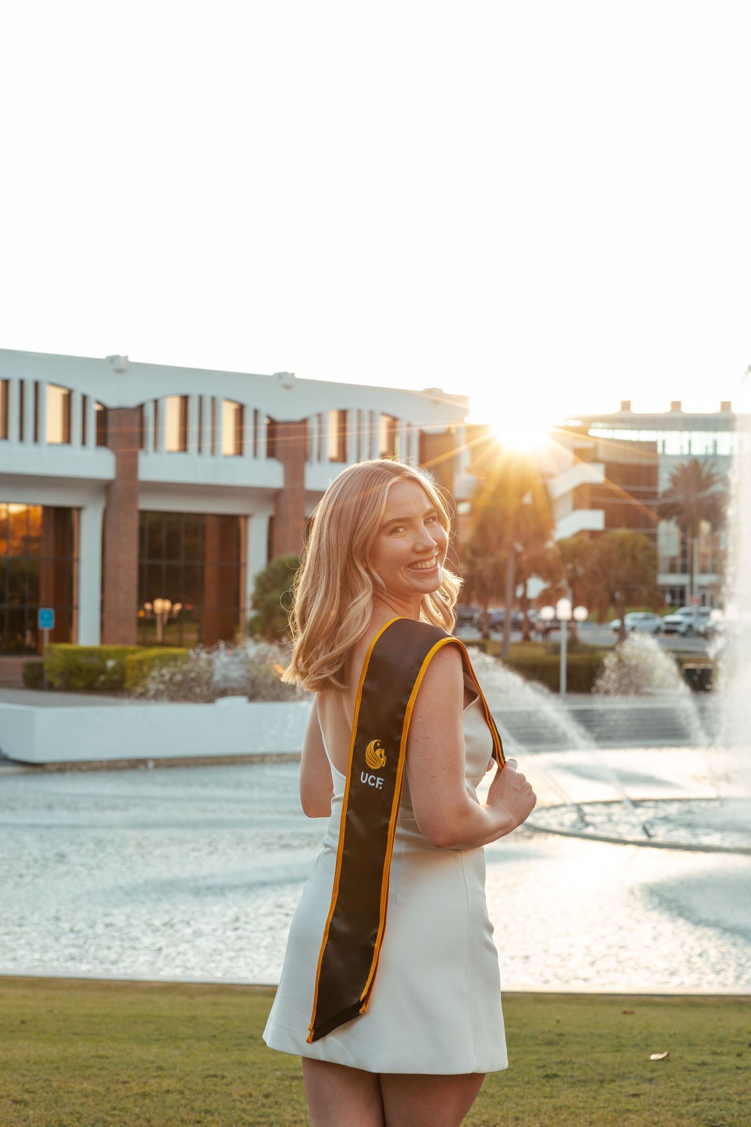 Graduate in white dress and stole poses near fountain at sunset during college graduation photoshoot.
