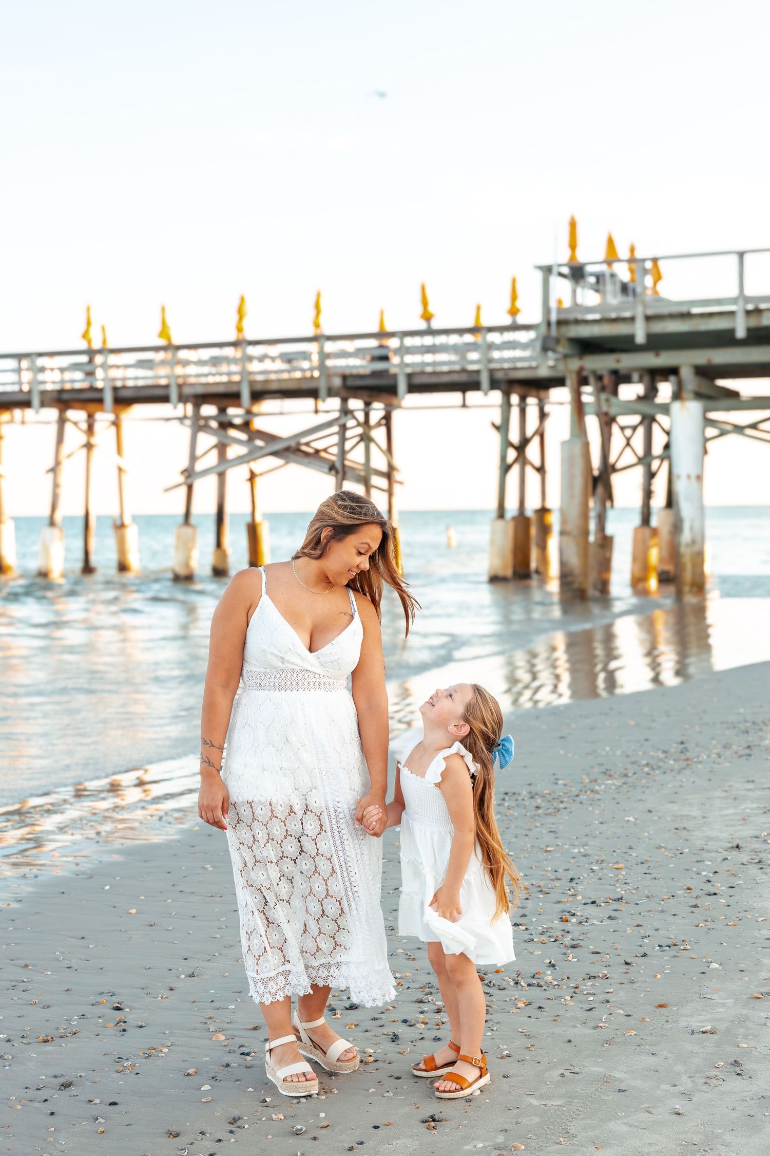 Two people in white lace dresses walk along a sandy beach near a wooden pier at sunset.