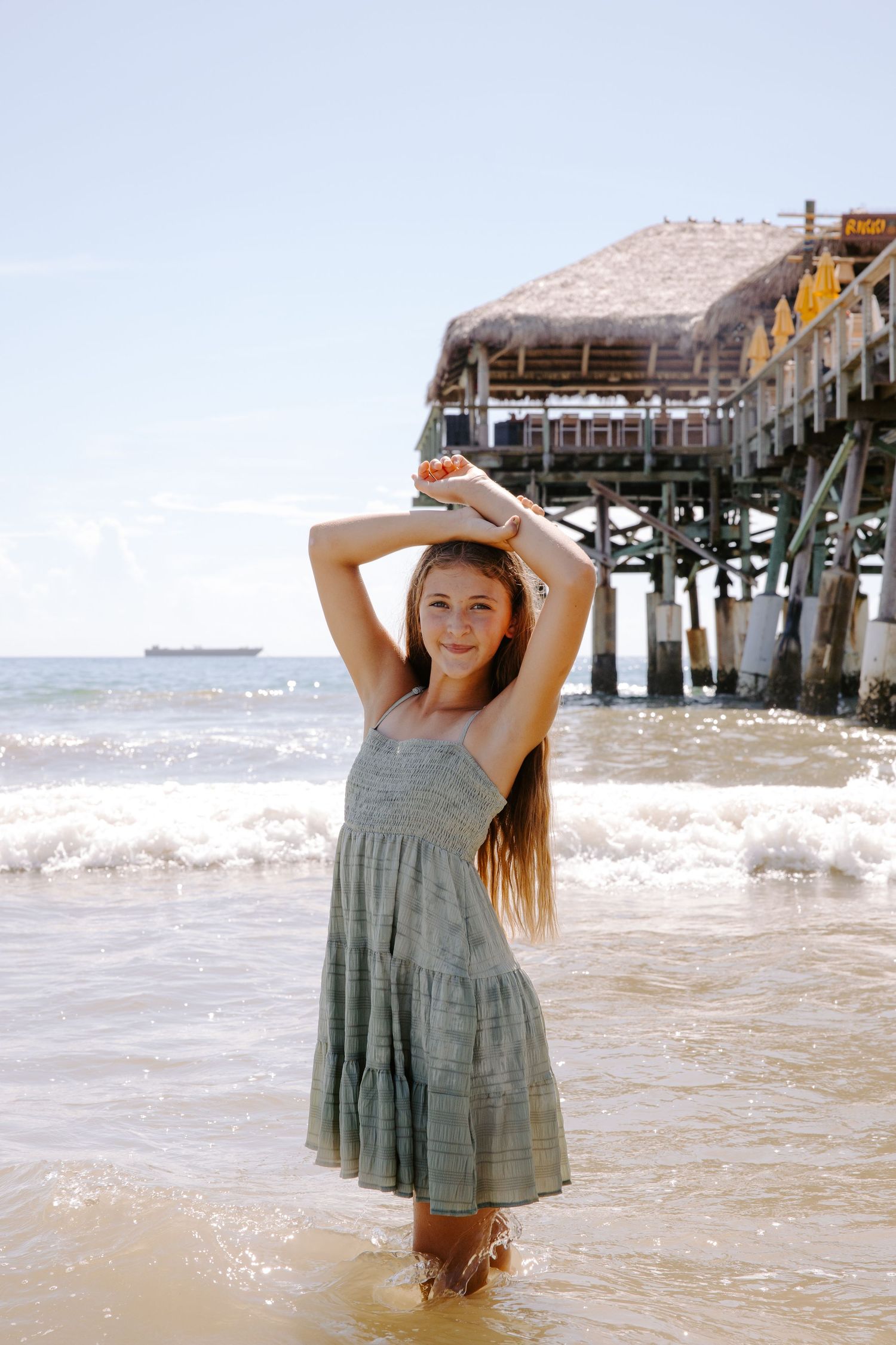 A young person in a grey dress plays in the ocean waves near a wooden pier.