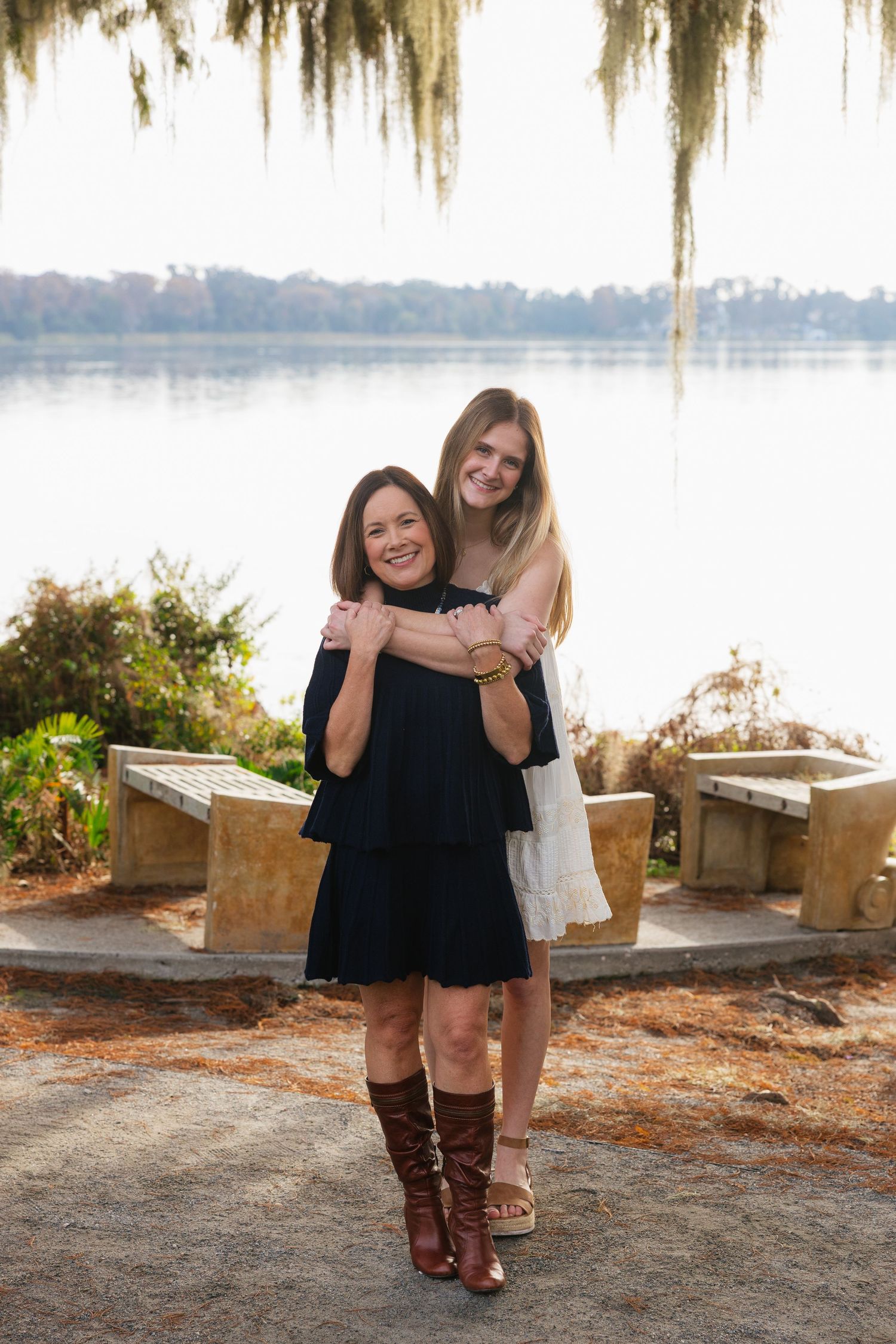 Two people in black dresses and brown boots pose together on a scenic waterfront path with Spanish moss overhead.