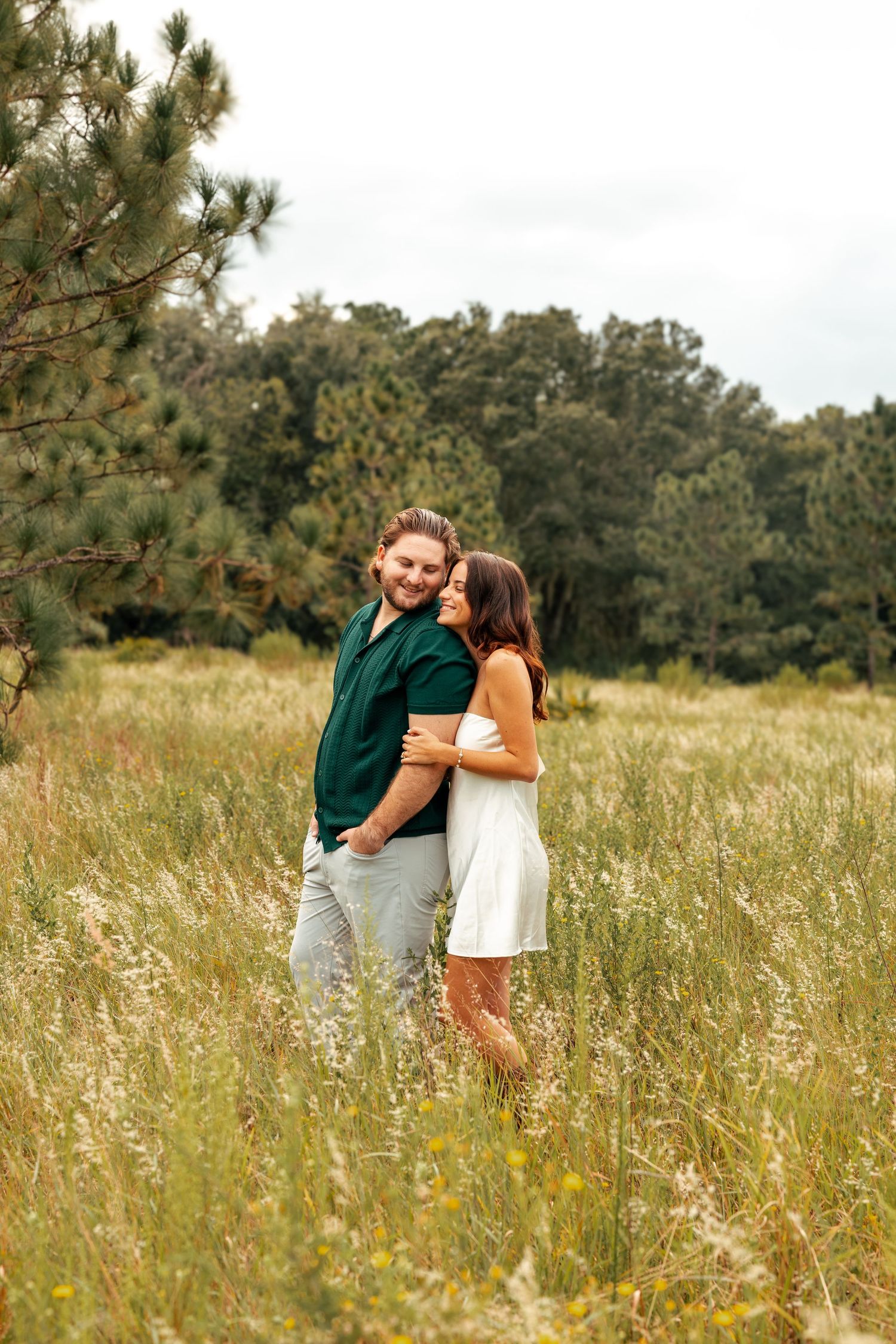 A couple shares an intimate moment in a sunlit field with tall grass and pine trees in the background.