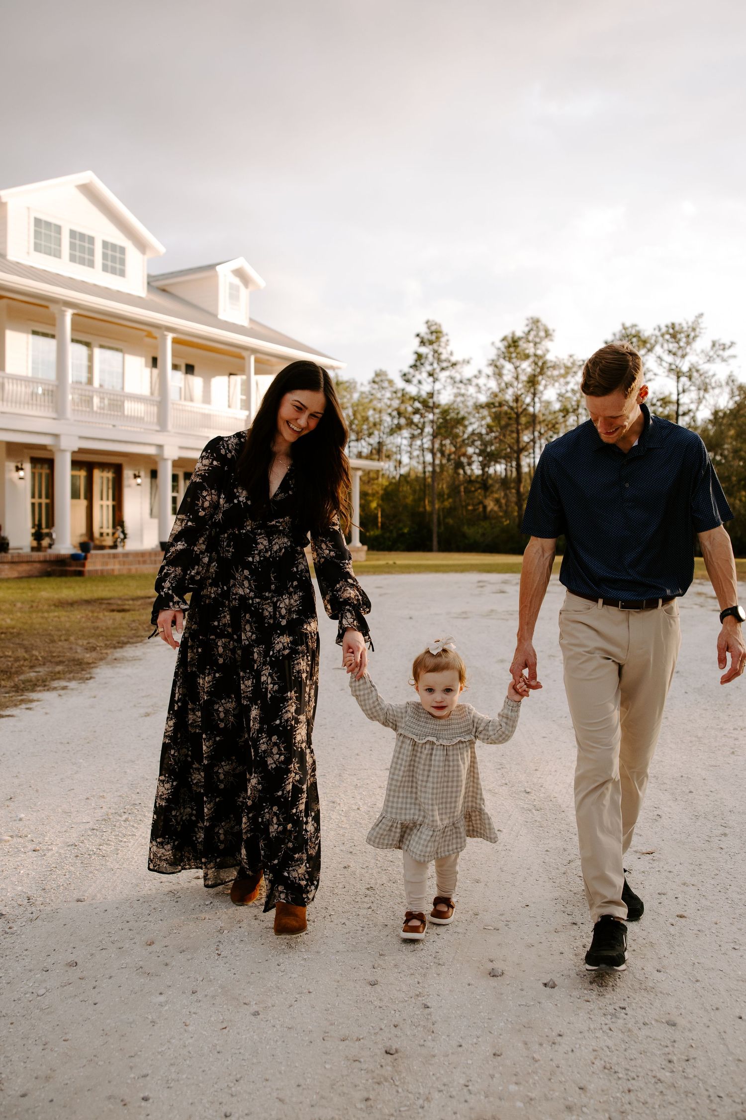 Family walking together on a sandy path with white farmhouse style building in background.