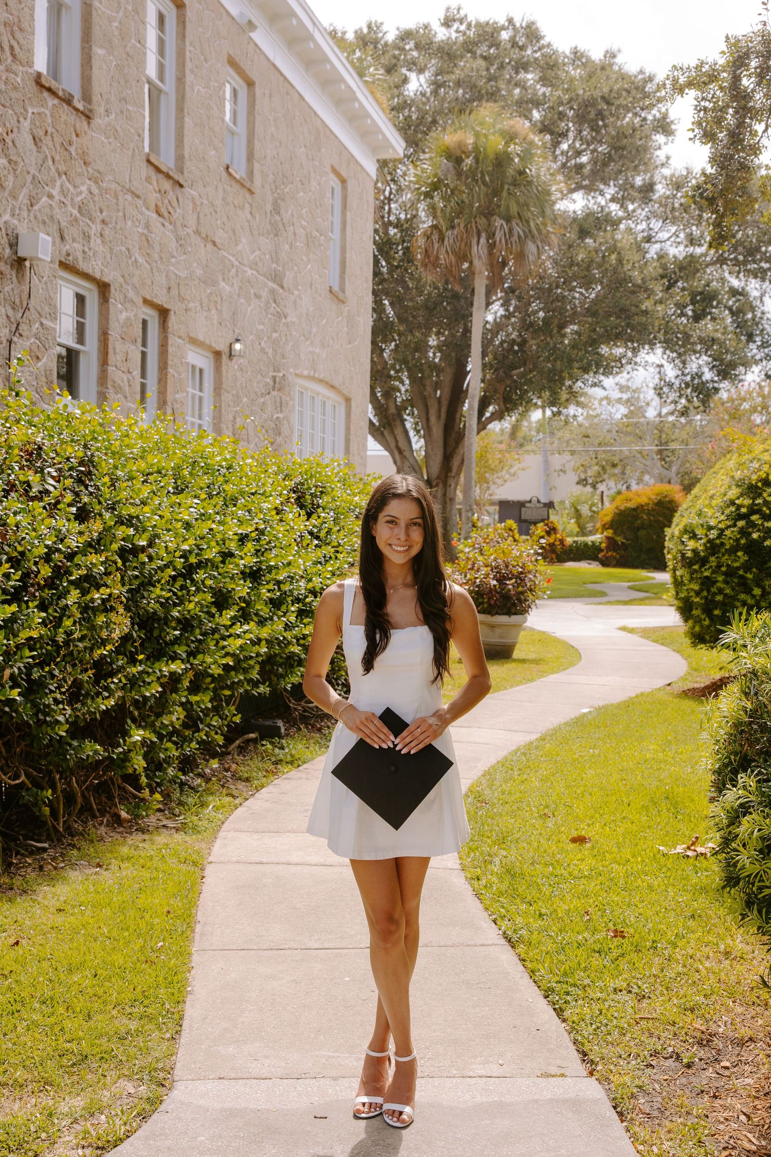 Student poses with diploma cover on a sunny campus sidewalk with buildings in background.