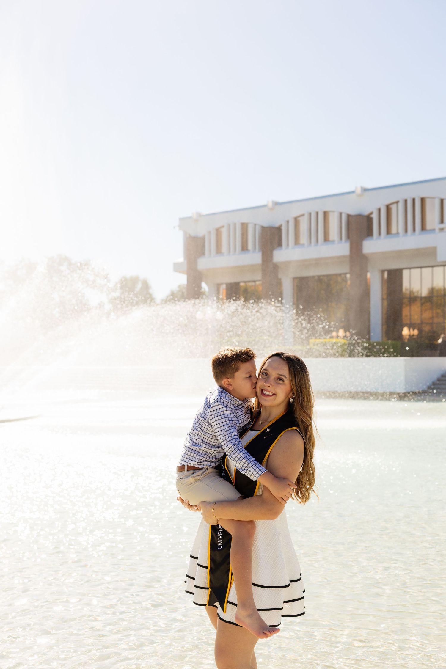 A romantic couple embraces and laughs together on a bright beach with a building in the background.
