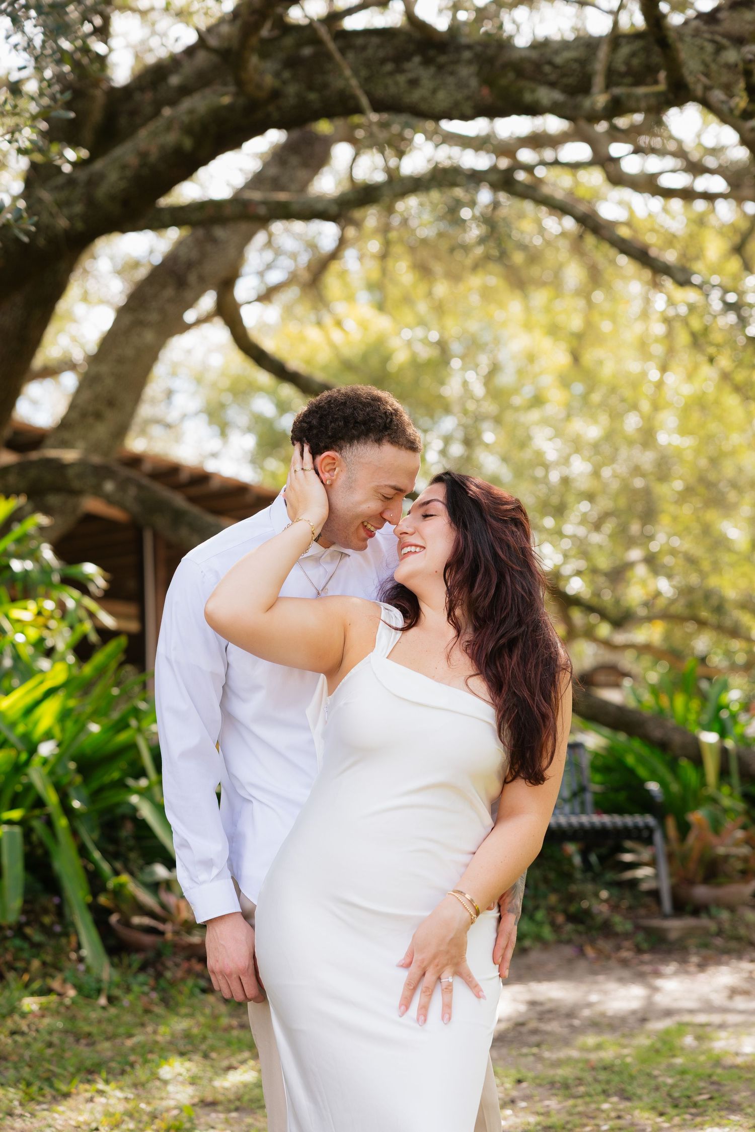 Couple embracing outdoors under large oak tree, woman in white dress smiling, man in white shirt, lush green garden background.