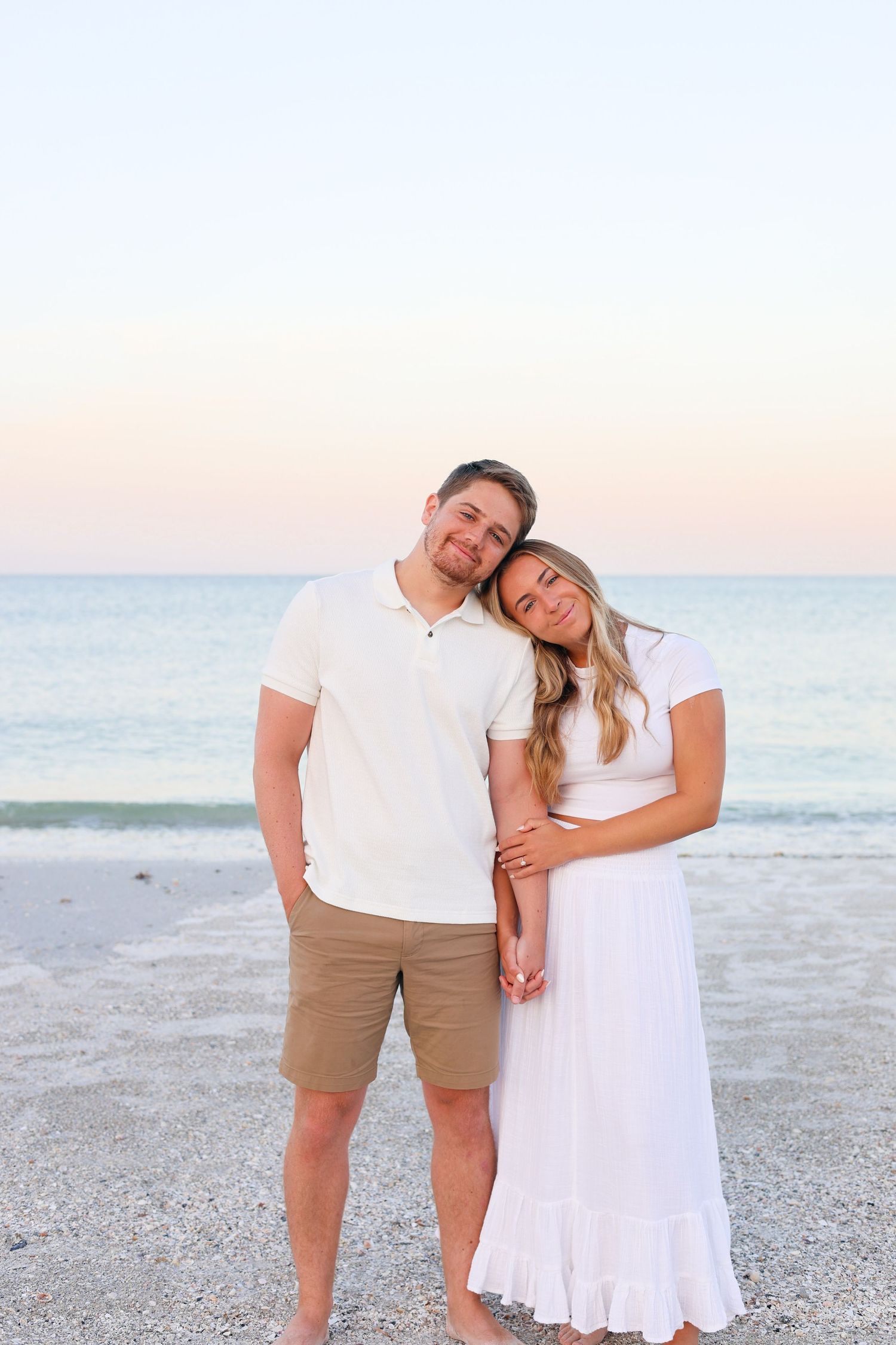 A couple in white outfits poses together on a serene beach at dusk.