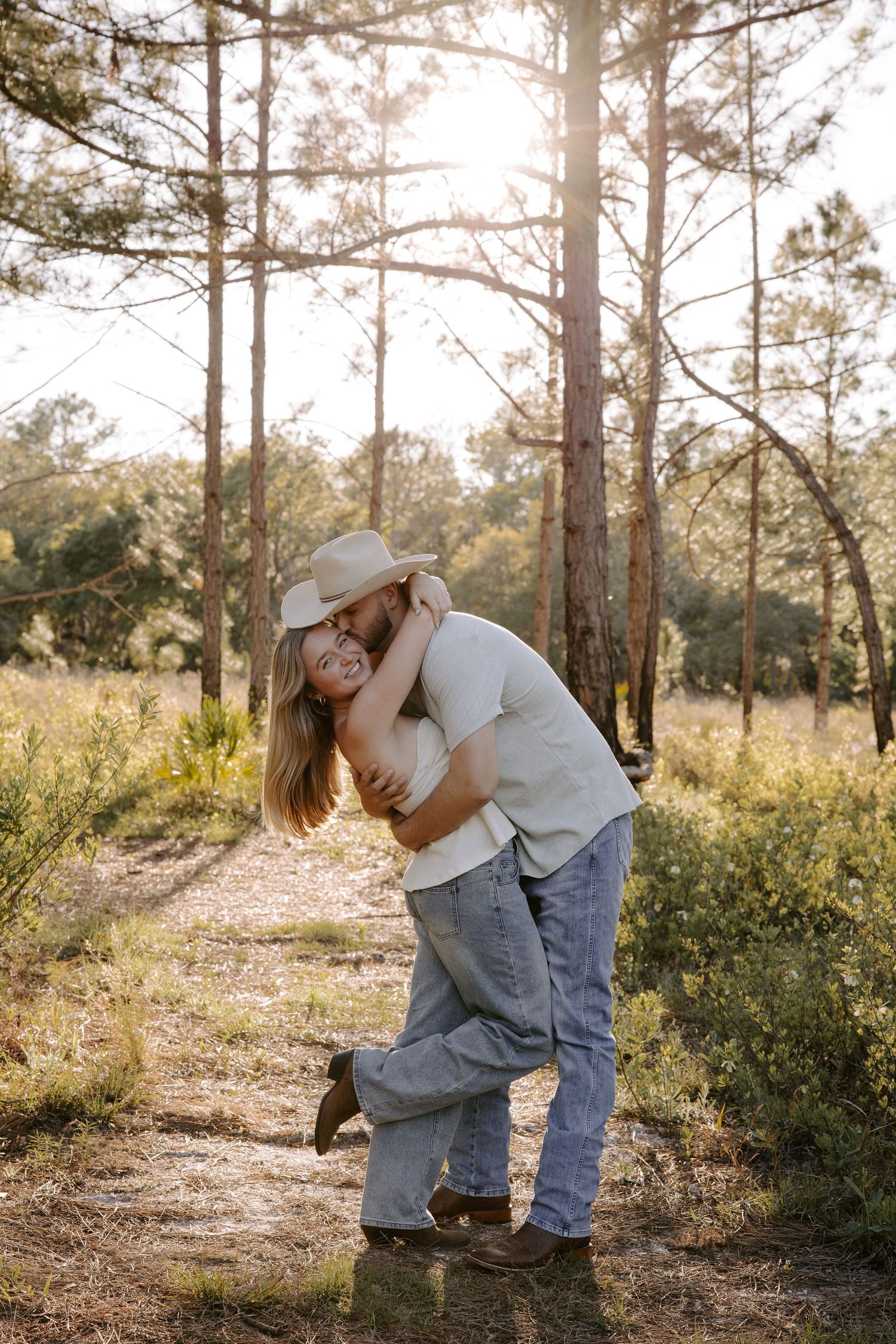 A couple embraces playfully among pine trees in a wooded setting.