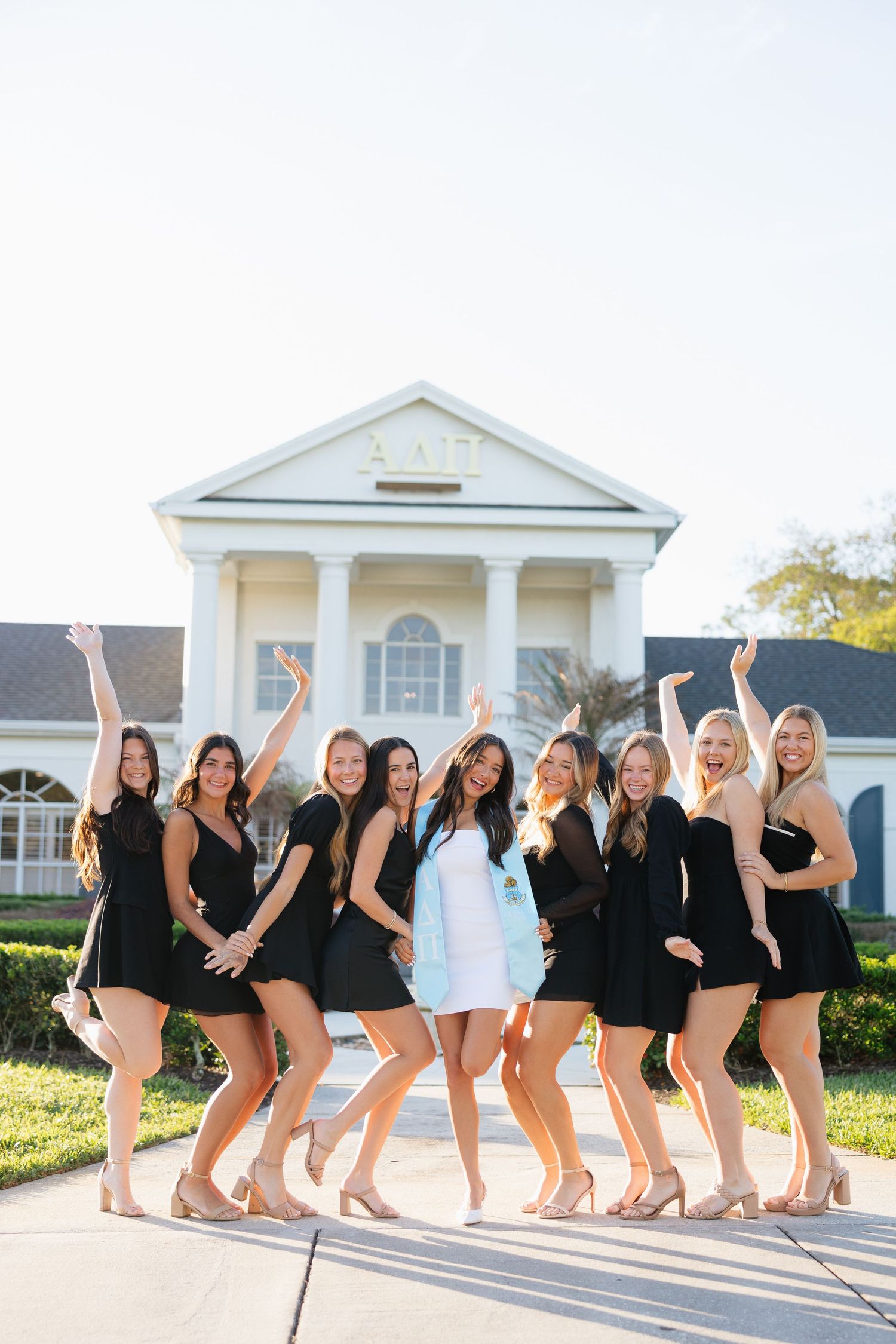 Group of women in black dresses posing in front of a sorority house on a sunny day.