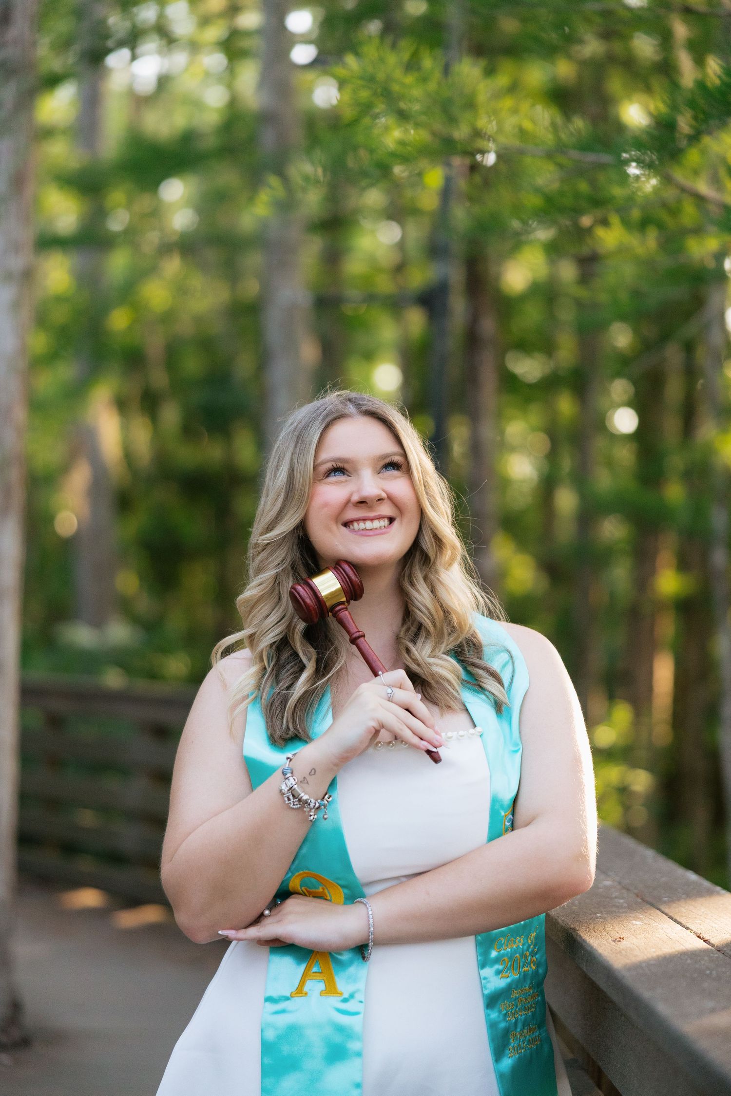 Smiling blonde woman in white dress with teal graduation sash holds a gavel outdoors on a wooden bridge surrounded by trees.