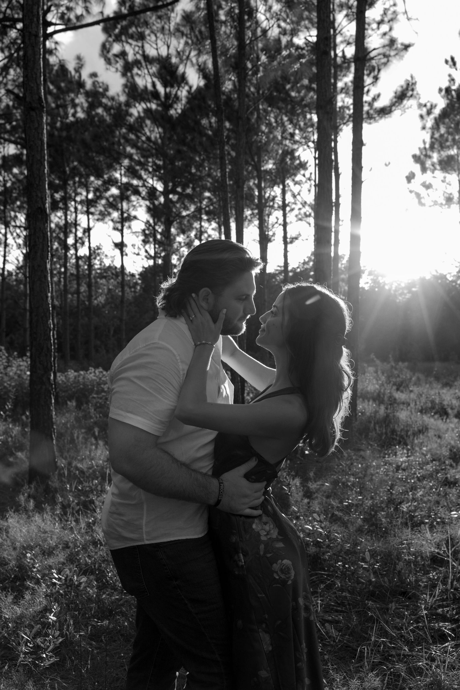 A black and white photo series shows two people in a tender romantic embrace among tall pine trees at sunset.