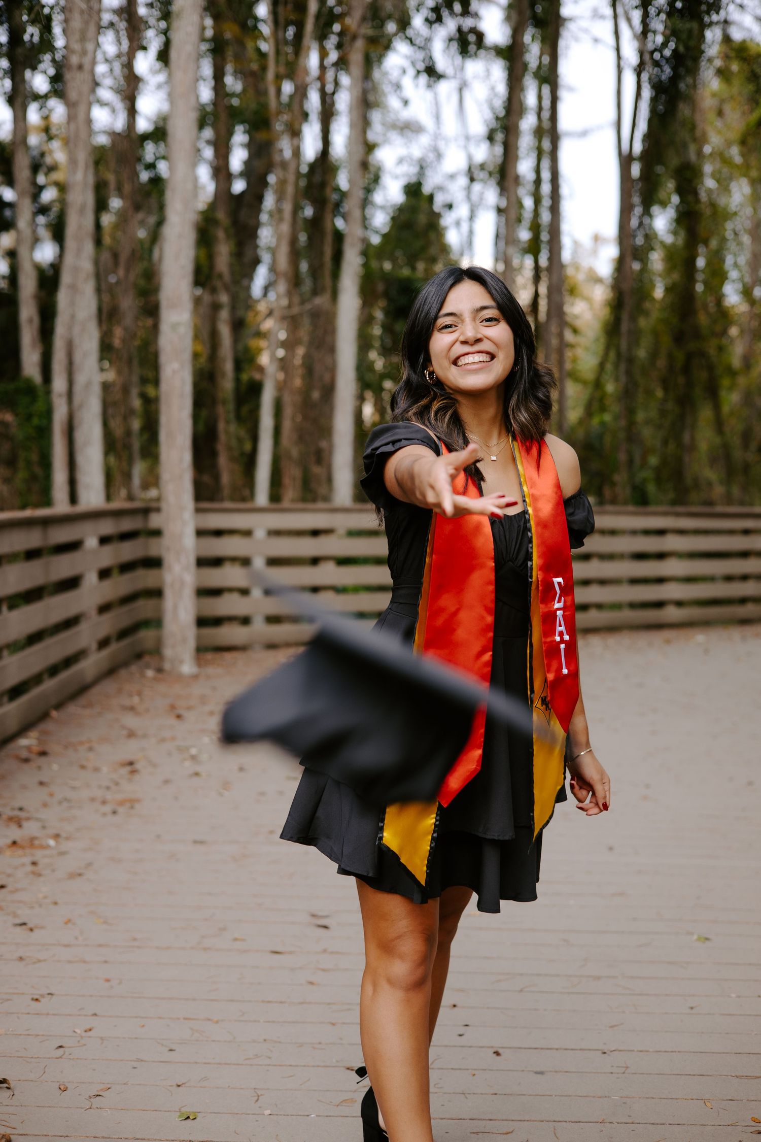Graduate in a vibrant red dress and black gown twirls on a boardwalk surrounded by pine trees.