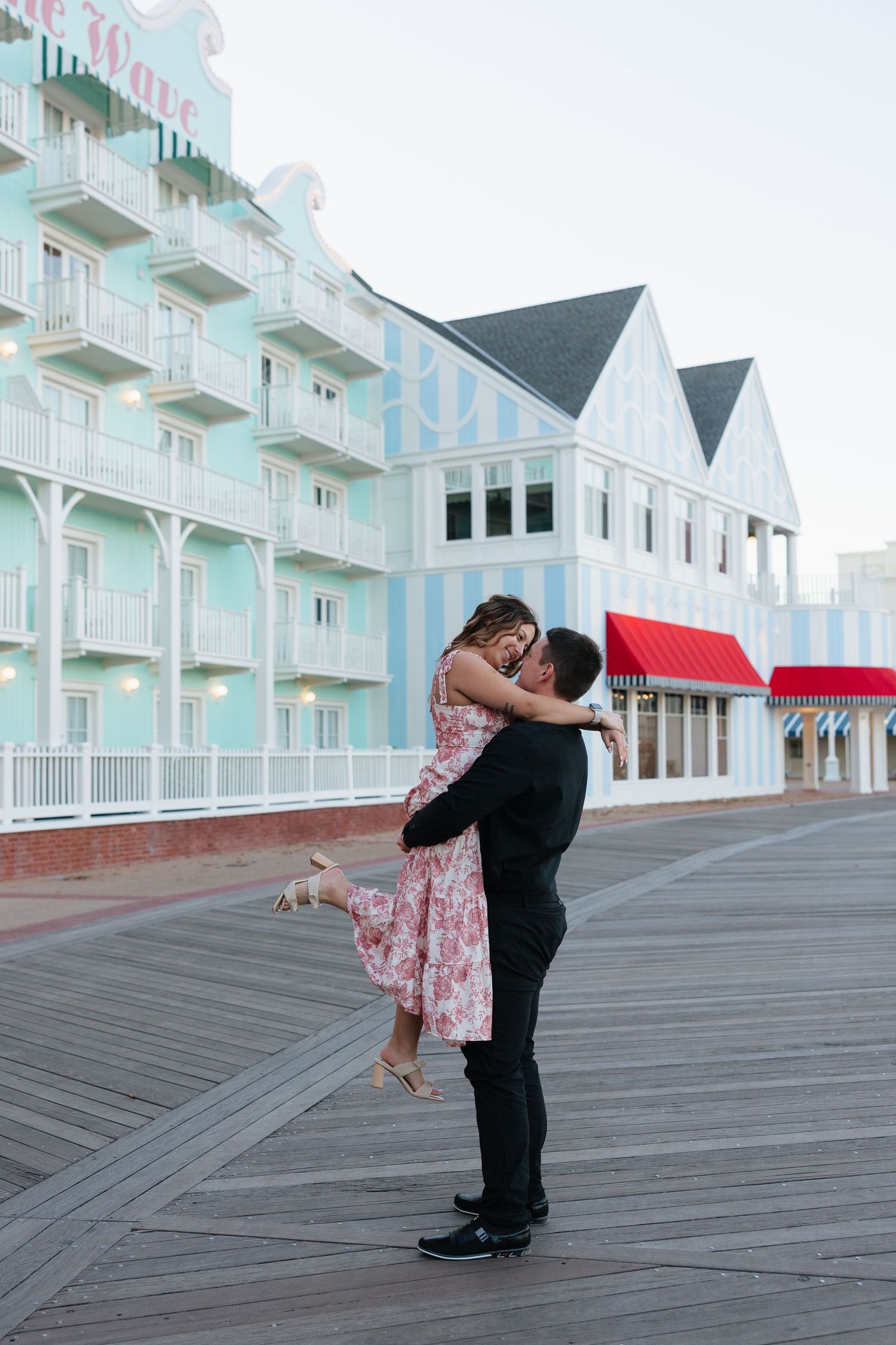 Couple embracing on a beachfront boardwalk with white Victorian-style buildings in the background.