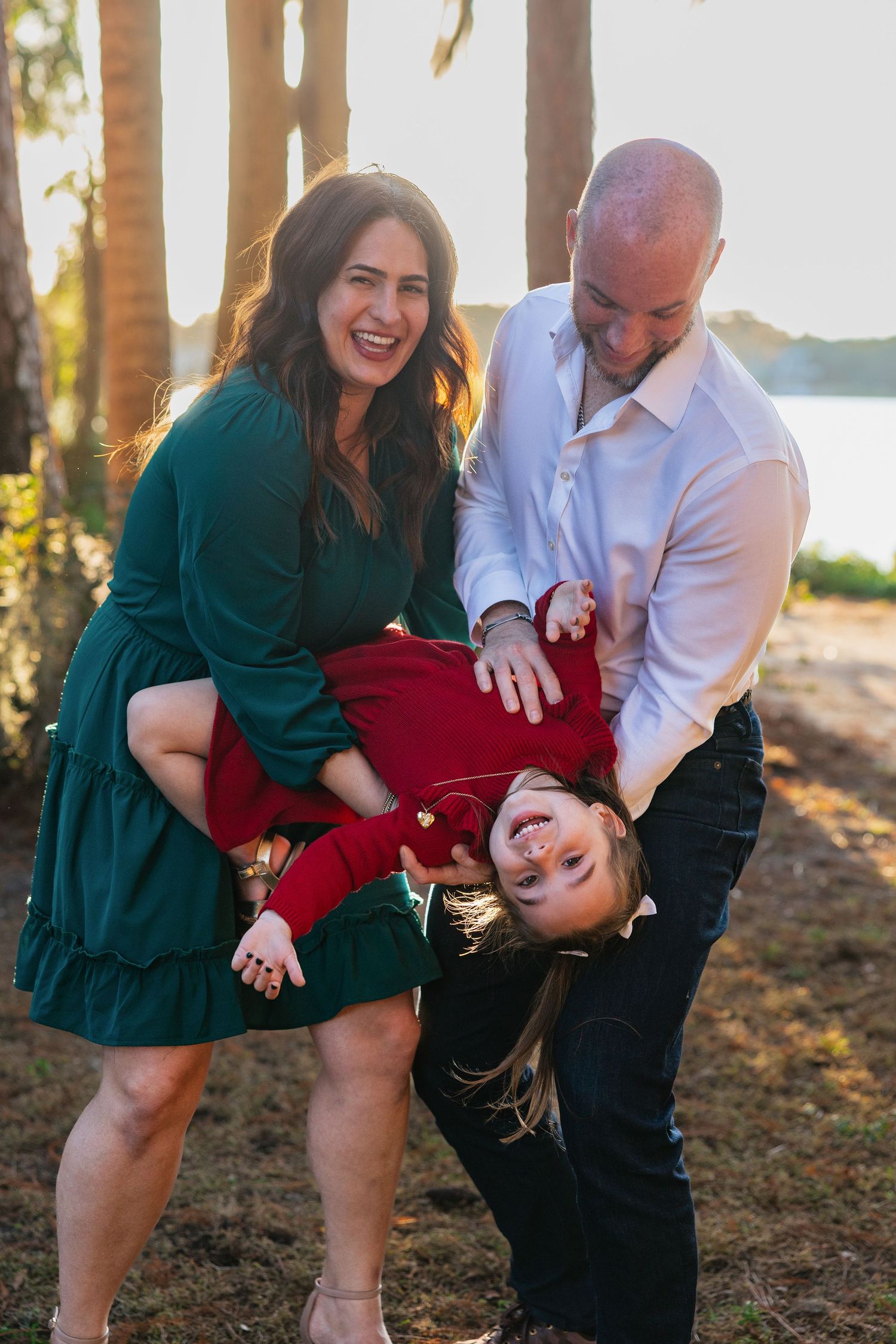 A family shares a playful moment together outdoors by a lake during golden hour, with trees in the background.