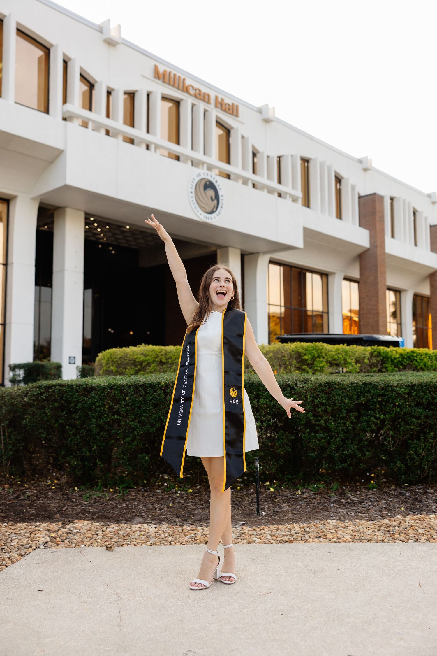 A person in a navy blazer and white dress stands joyfully with arms raised outside a white building.