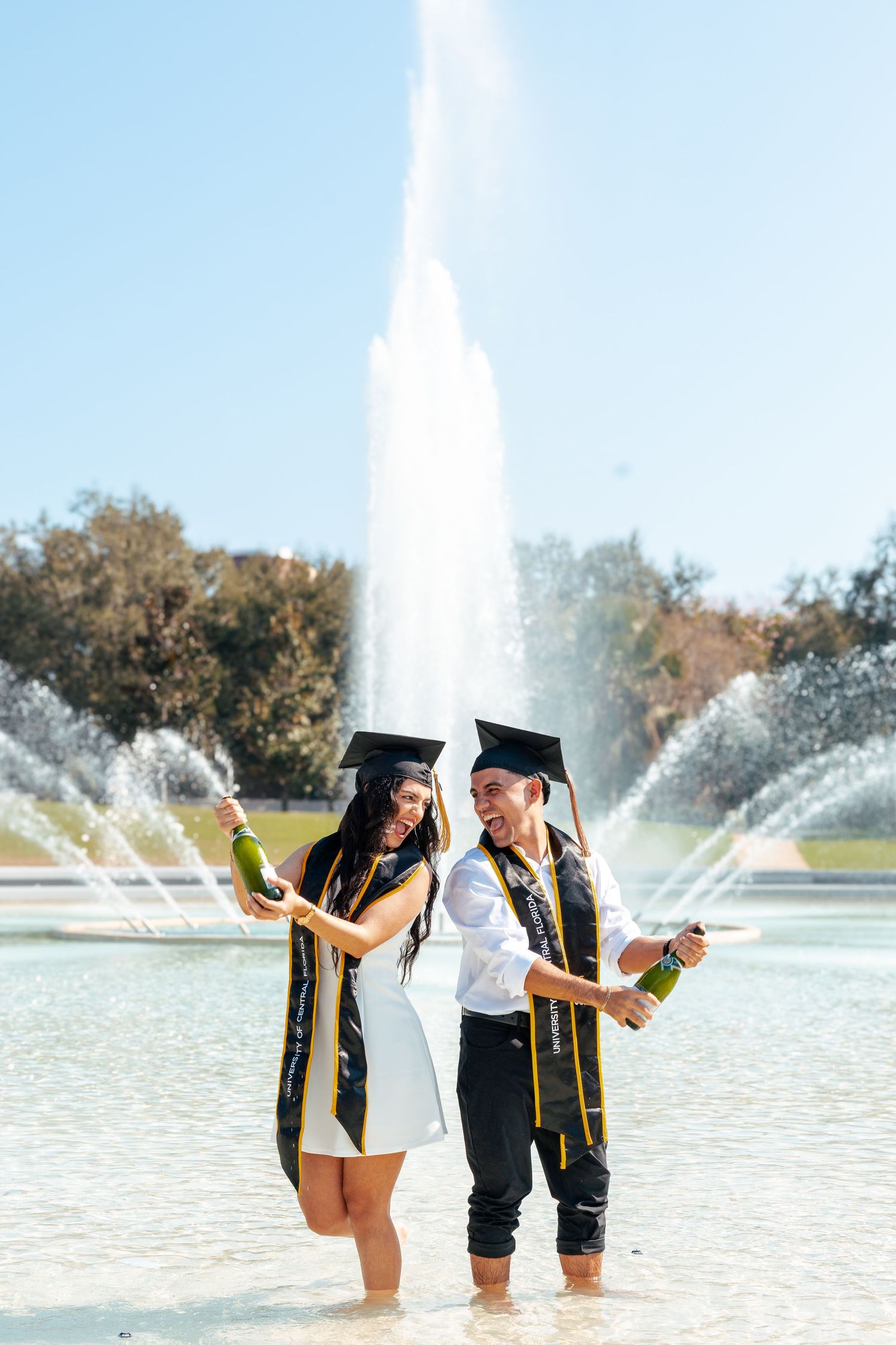 Graduates in caps and gowns celebrate by popping champagne bottles near a fountain on their graduation day.
