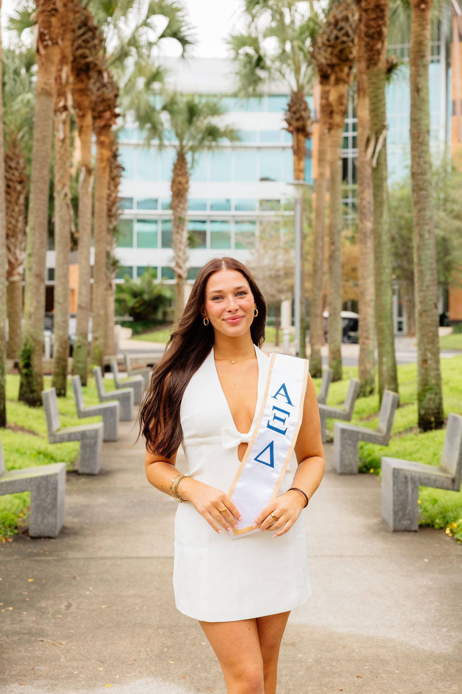A person in white dress stands on a palm tree-lined walkway at a university campus.