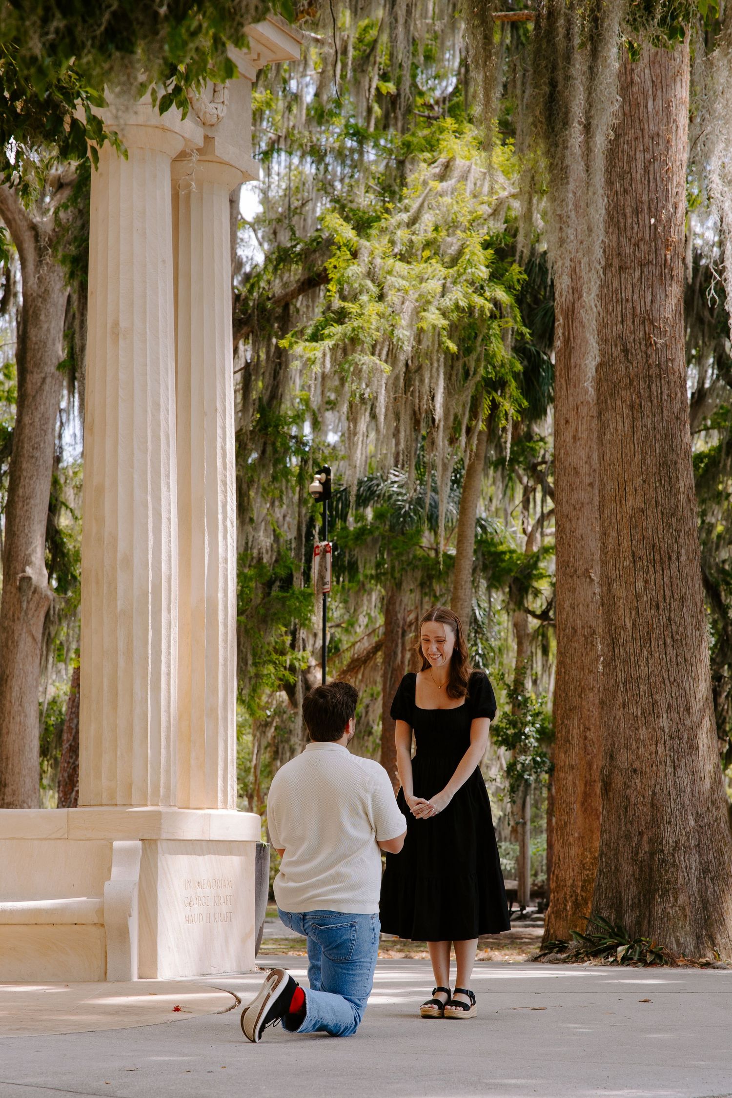 A couple sits and stands among white columns and Spanish moss draped trees in a serene outdoor setting.