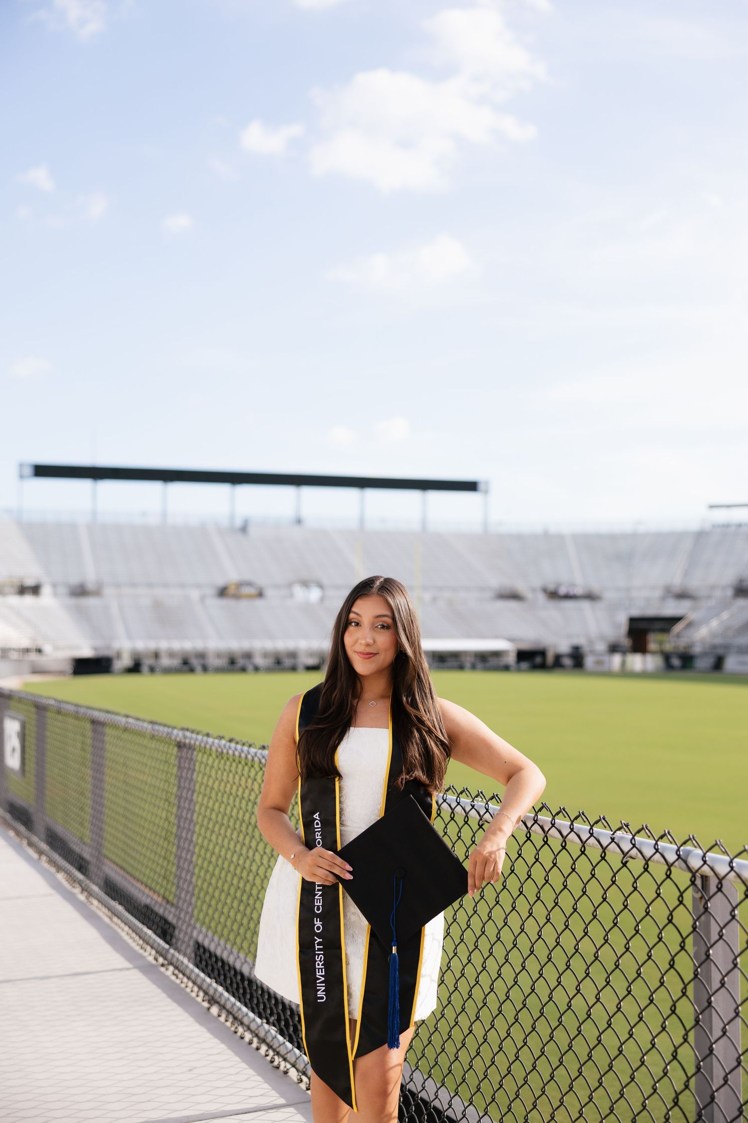 A young woman in graduation attire poses at a stadium, wearing a black cap and gown with a gold University sash.