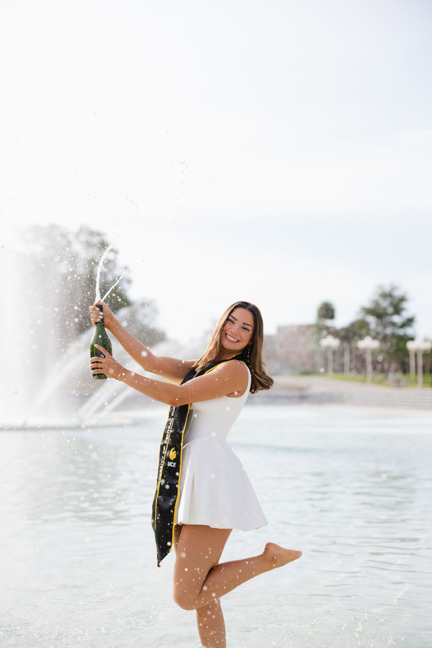 A smiling young woman in a white dress pops a champagne bottle near a fountain outdoors.