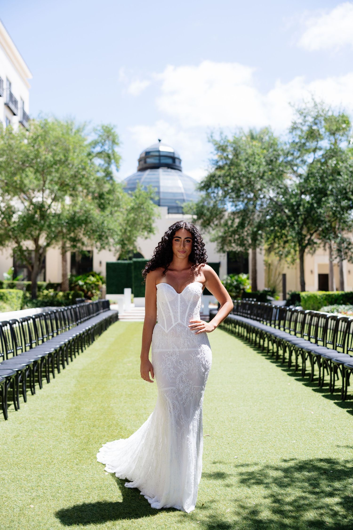 Bride in white lace mermaid gown stands between rows of black chairs in an outdoor garden ceremony venue.