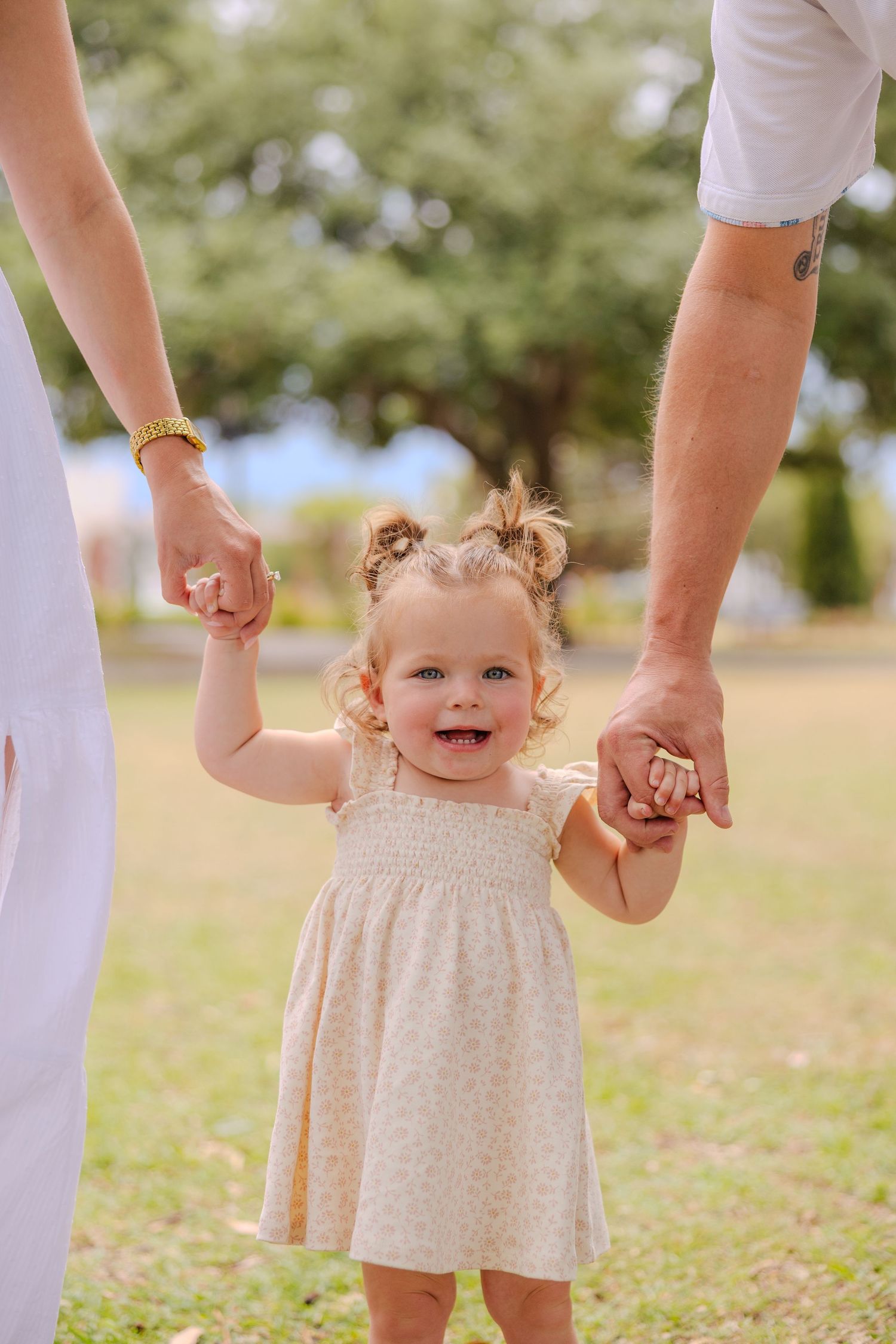 Smiling toddler girl in floral dress holds hands with parents while walking outdoors in a sunny green park.