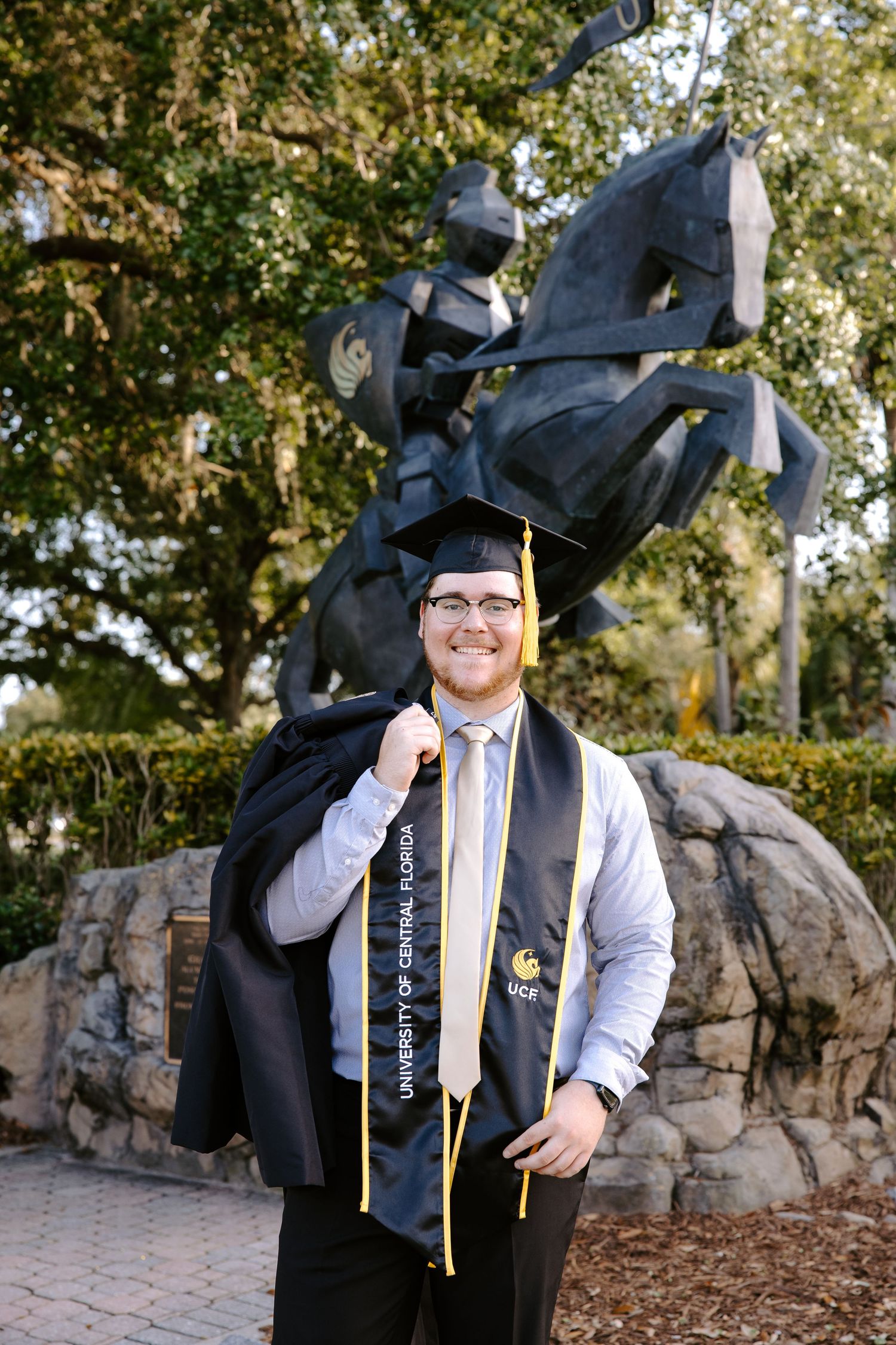 Graduate in cap and gown poses near stone wall with UCF tassel and honors cords.