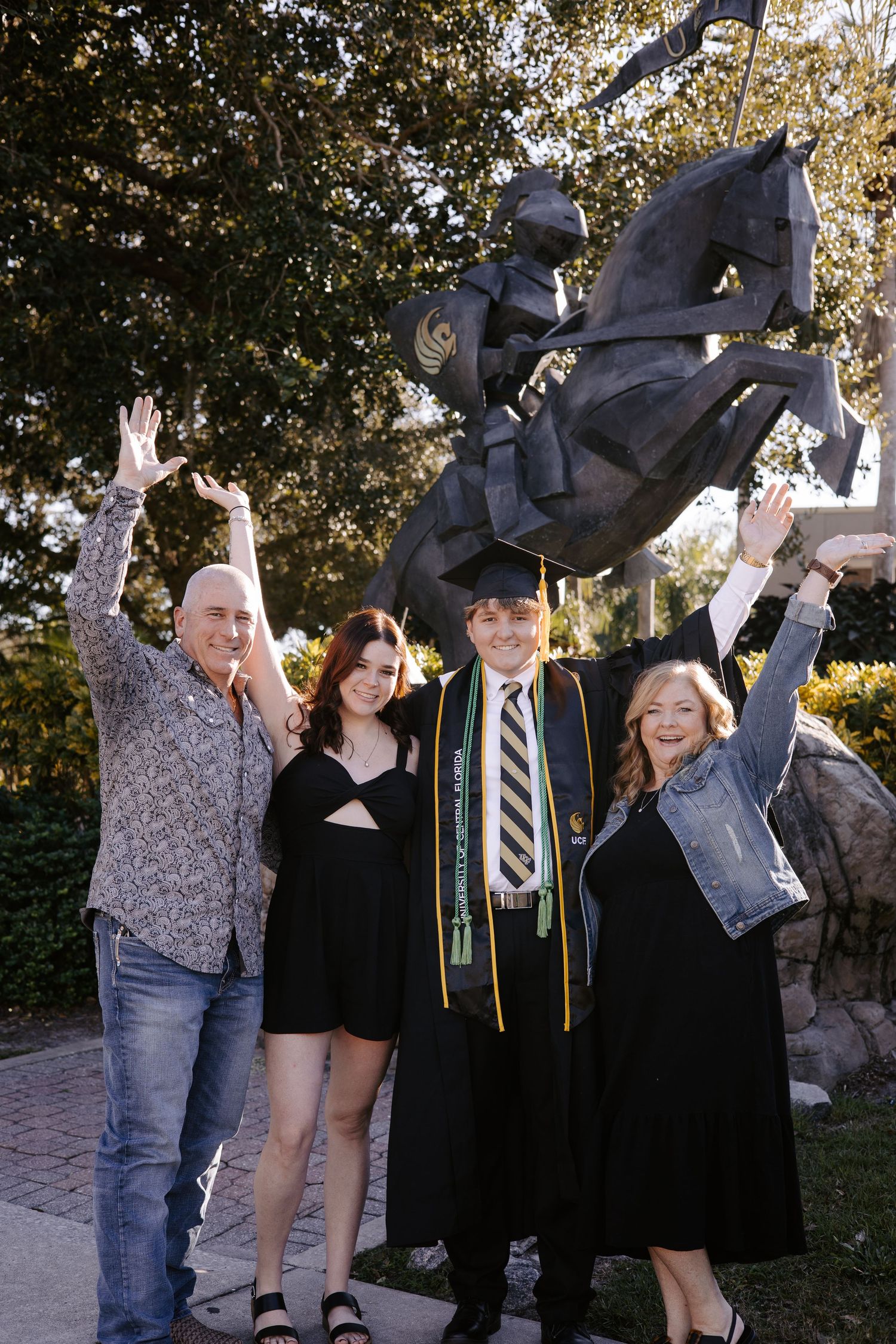 Group celebrates in front of a stone statue wearing graduation attire and casual clothes.