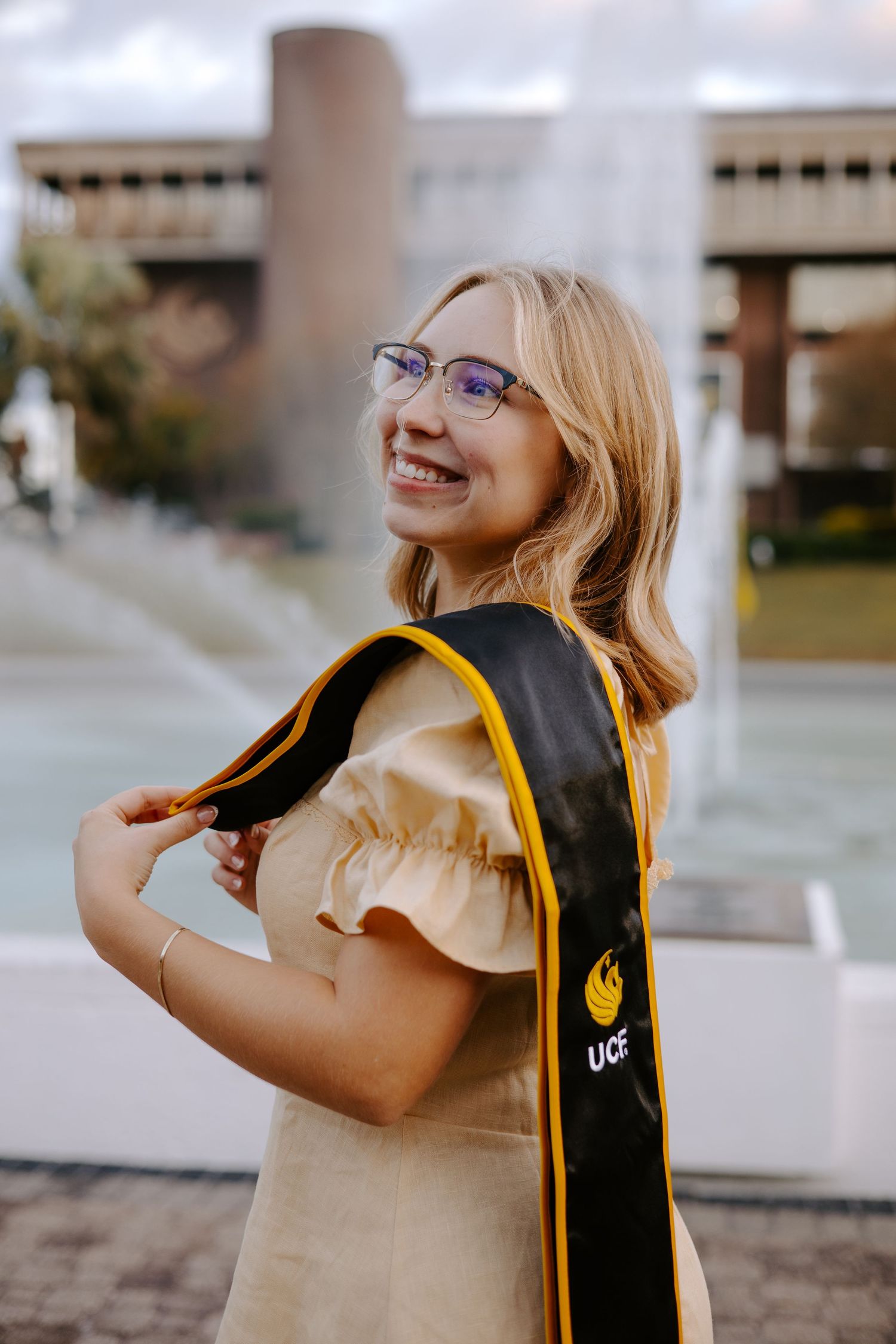 Graduate in beige dress holds UCF graduation stole while posing outdoors.