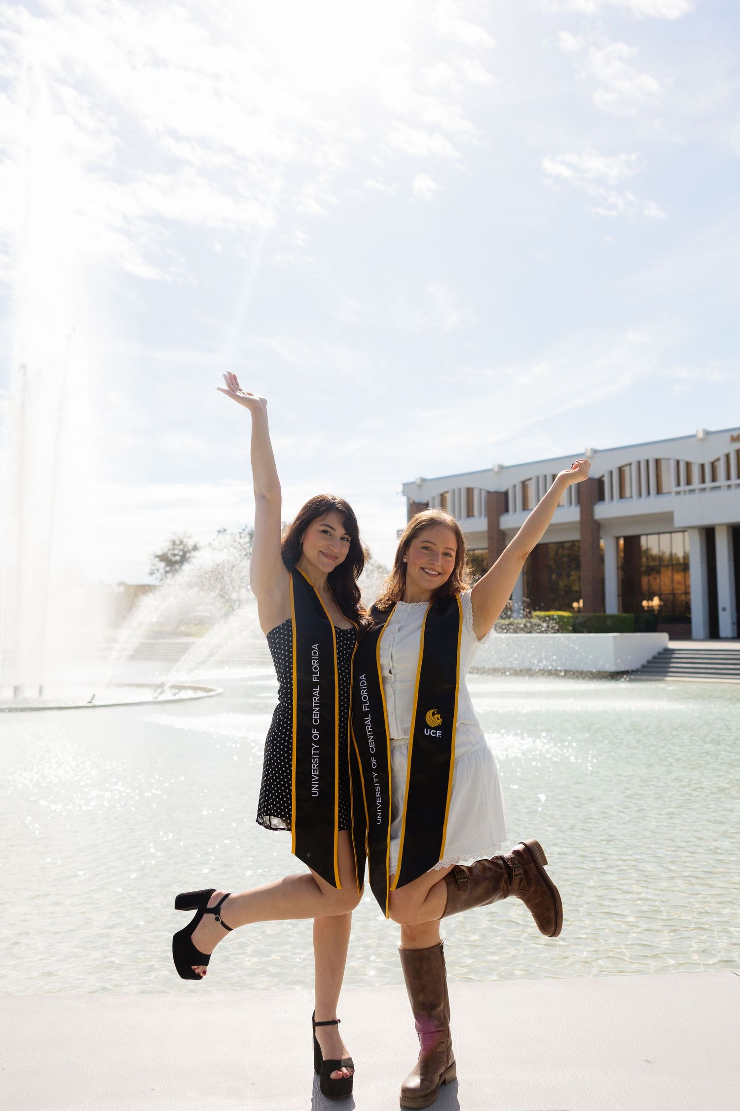 Two people in black graduation gowns pose playfully in front of a fountain on a sunny day at a university campus.