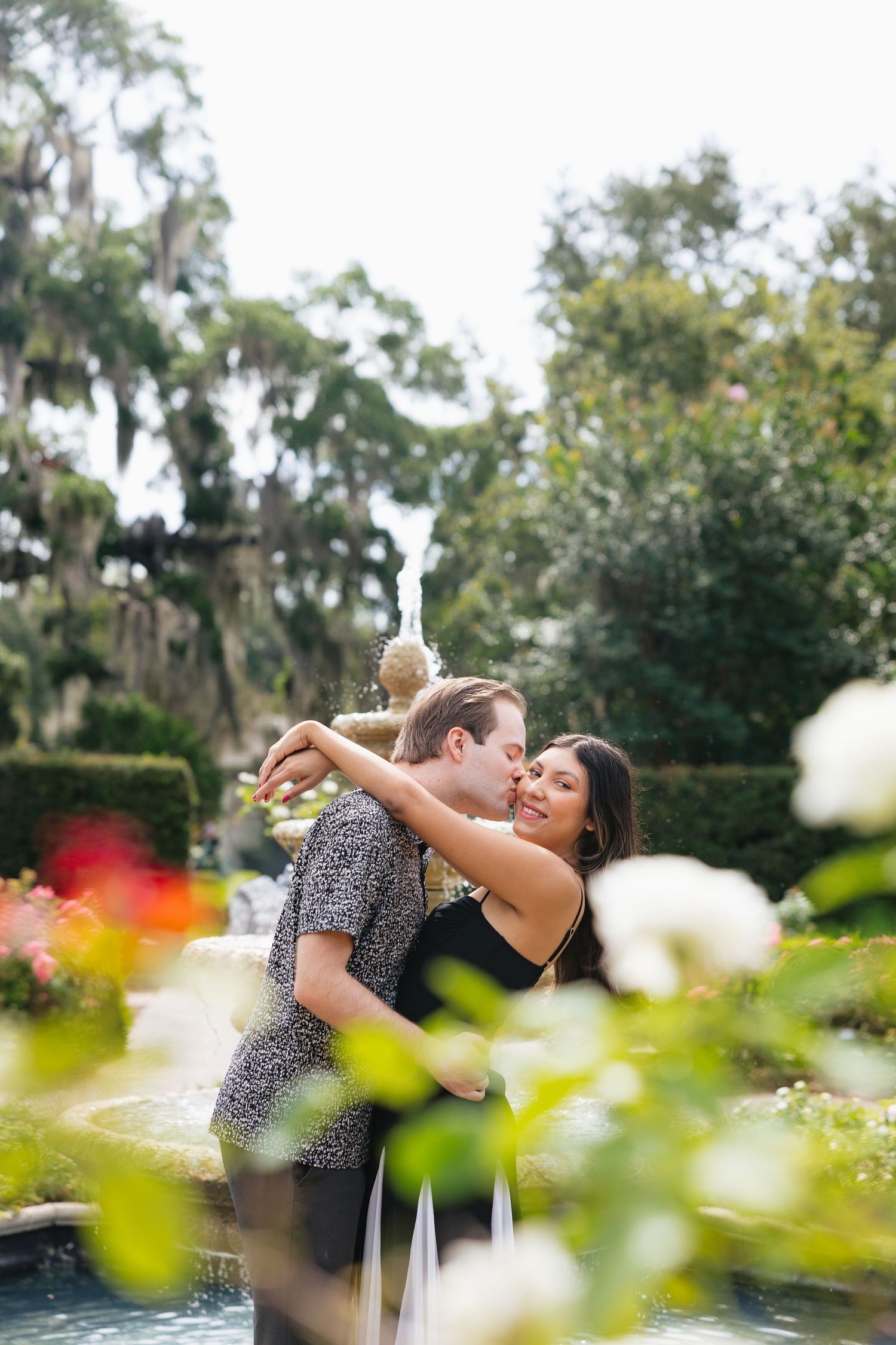 Couple embracing and kissing near a garden fountain surrounded by lush greenery and colorful flowers.