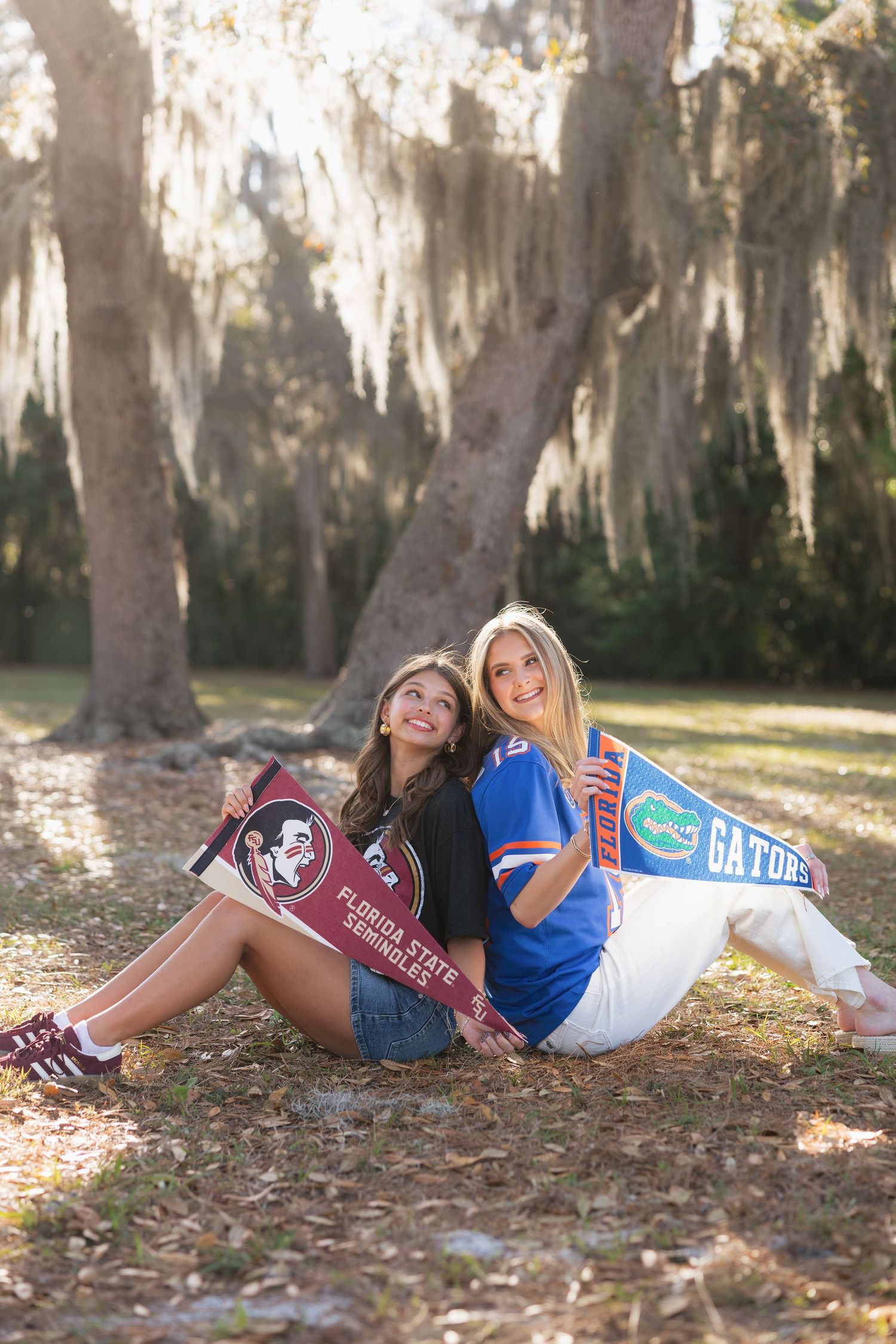 Two college fans sit outdoors holding Florida State Seminoles and Florida Gators pennants, smiling under Spanish moss-draped trees.