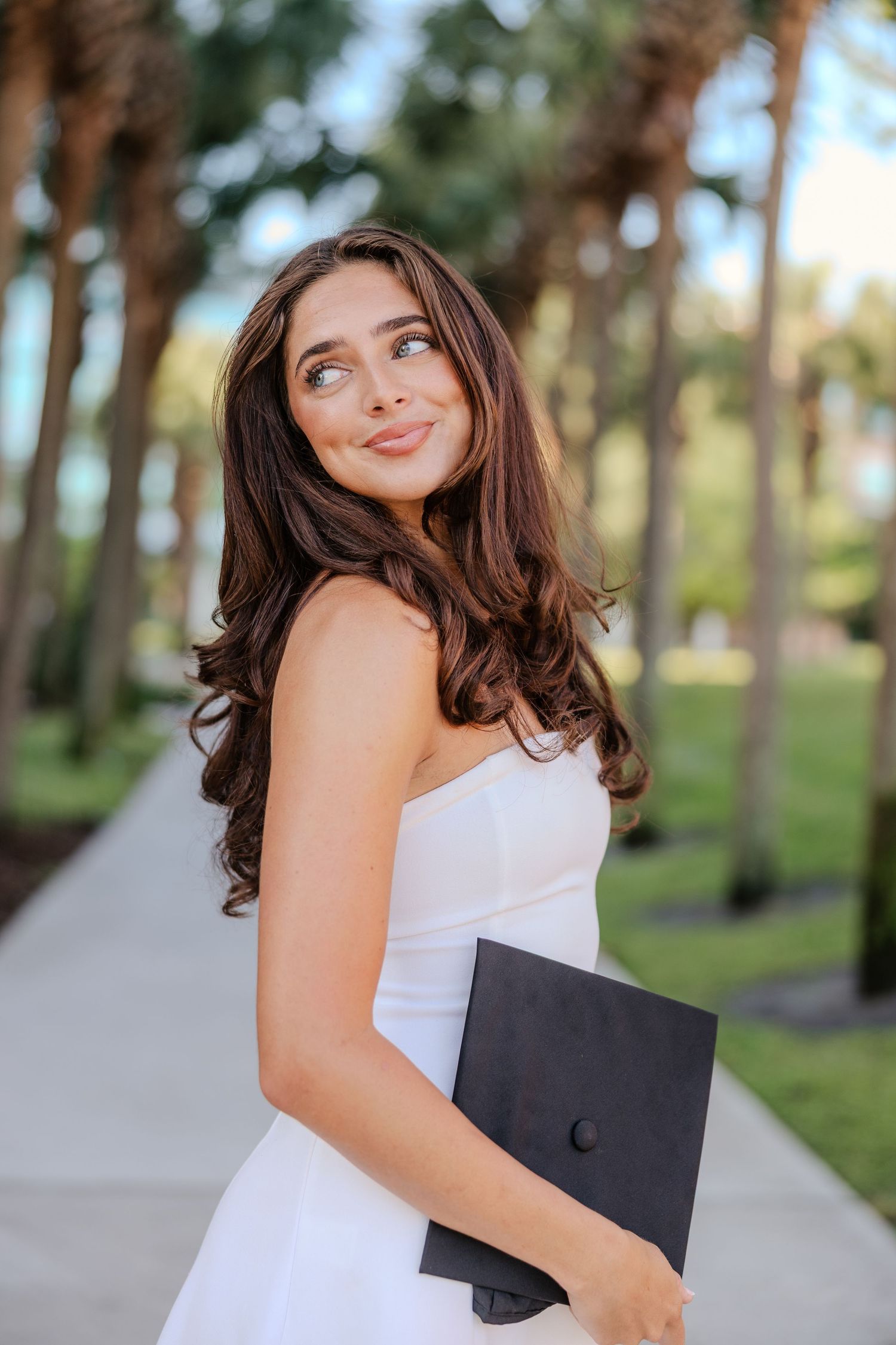 Young woman in white strapless dress holding graduation portfolio outdoors among palm trees, smiling upward.
