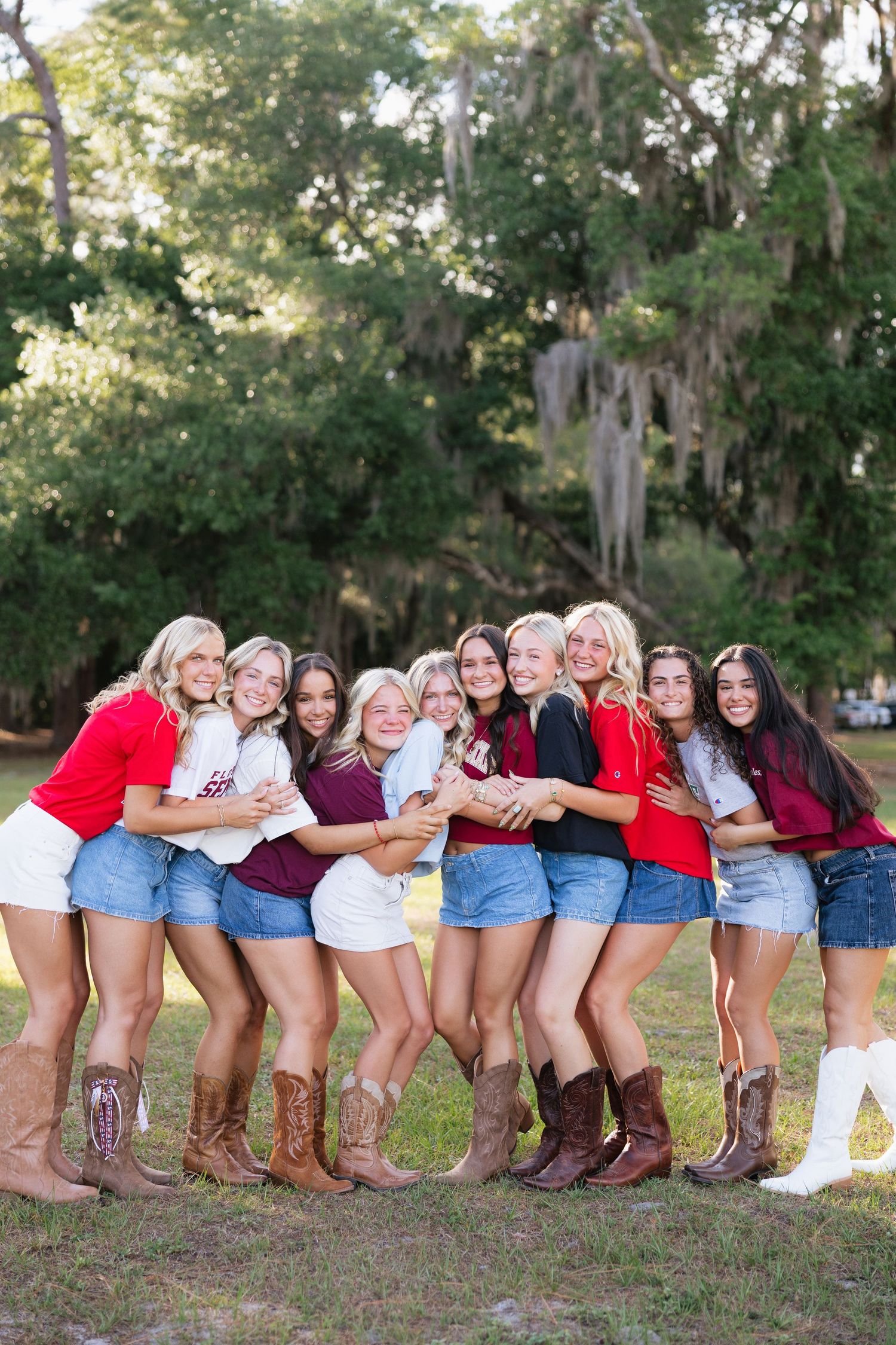 Group of young women in denim shorts and cowboy boots posing together outdoors under Spanish moss trees.