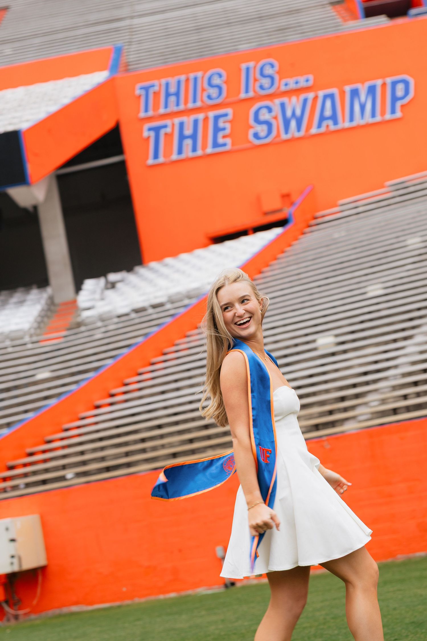 Smiling female graduate in white dress holding blue sash at The Swamp stadium, University of Florida.