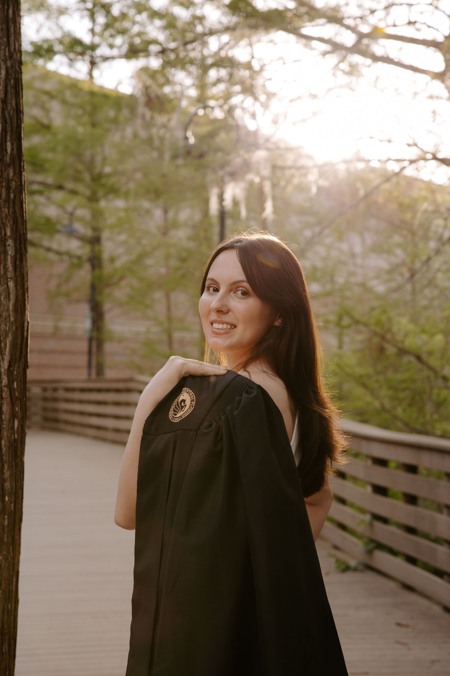 Graduate in black graduation gown poses on wooden boardwalk in forest.