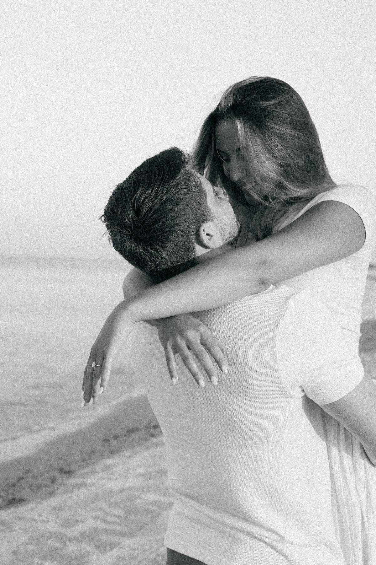 Couple in white clothing sharing intimate embrace on beach in black and white photography.