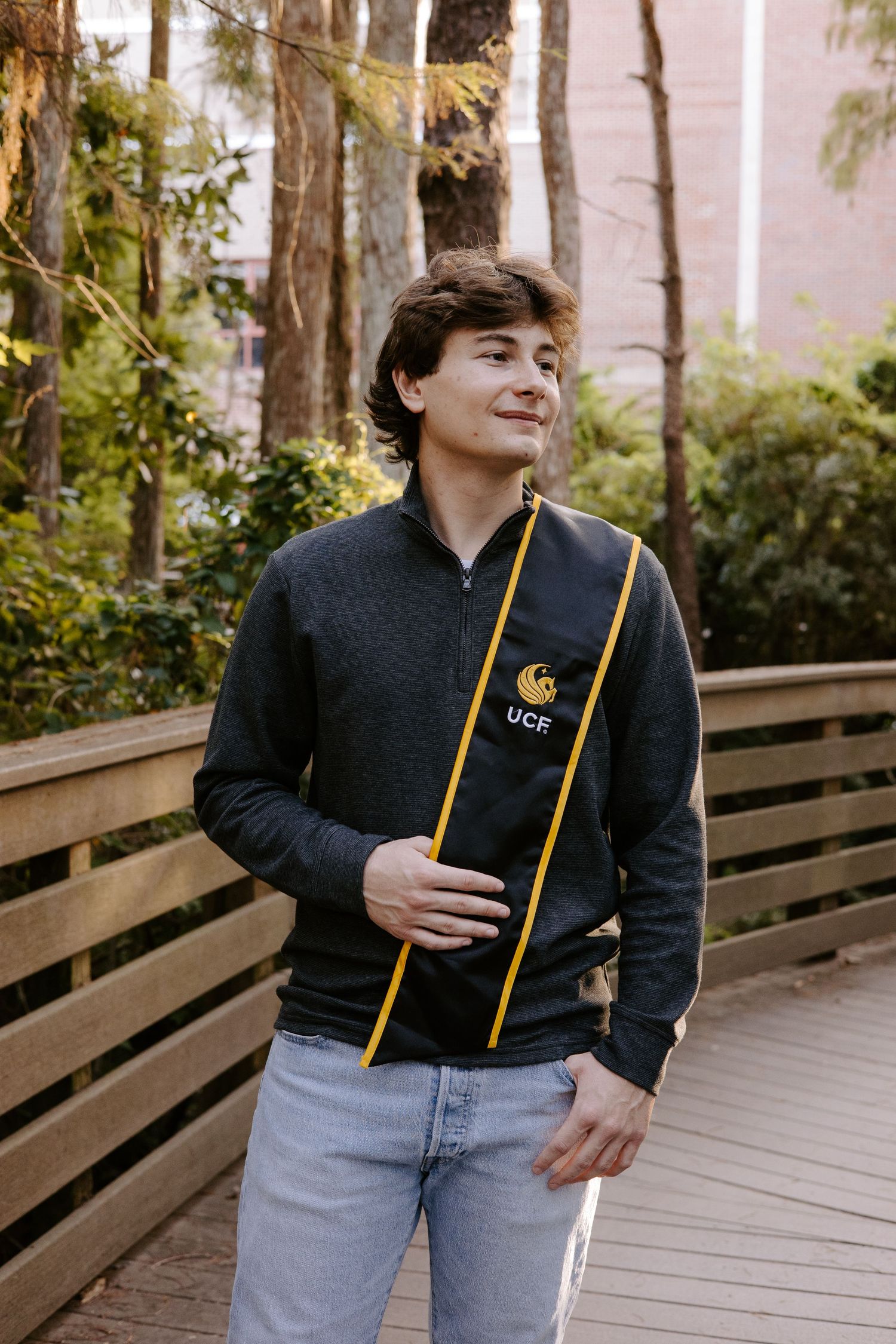Graduate in a black UCF hoodie and jeans poses on a wooden boardwalk surrounded by trees.