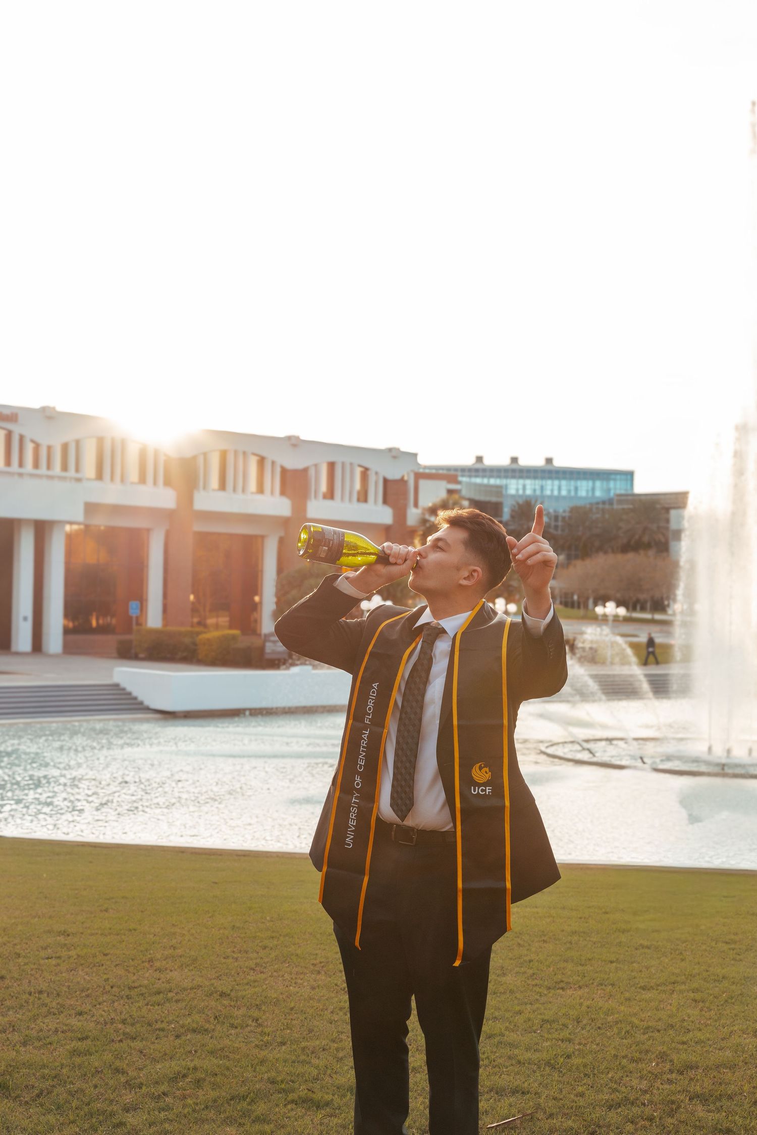 Graduate in black and gold academic regalia poses with arms outstretched on a grassy lawn near campus buildings.