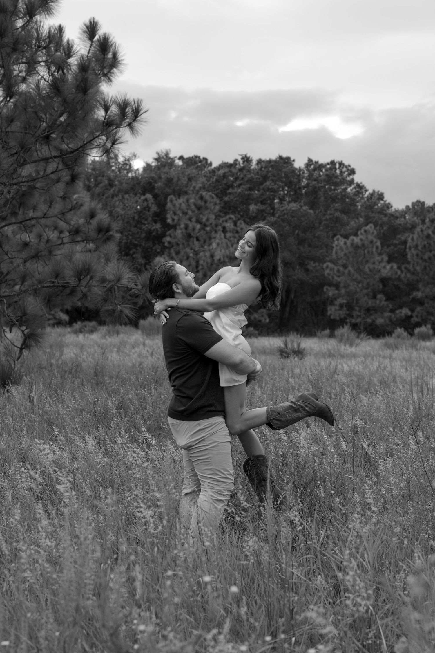 A romantic black and white moment captured in a field with pine trees in the background.