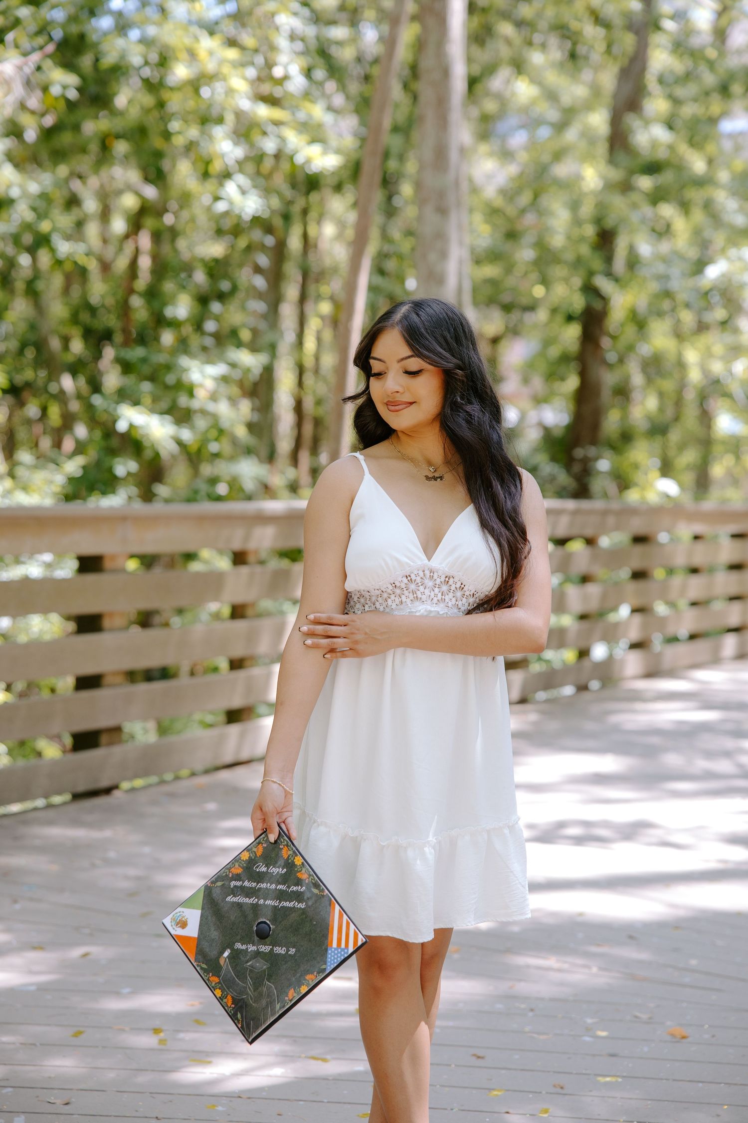 Graduate in white dress twirls with decorated graduation cap on wooden boardwalk.