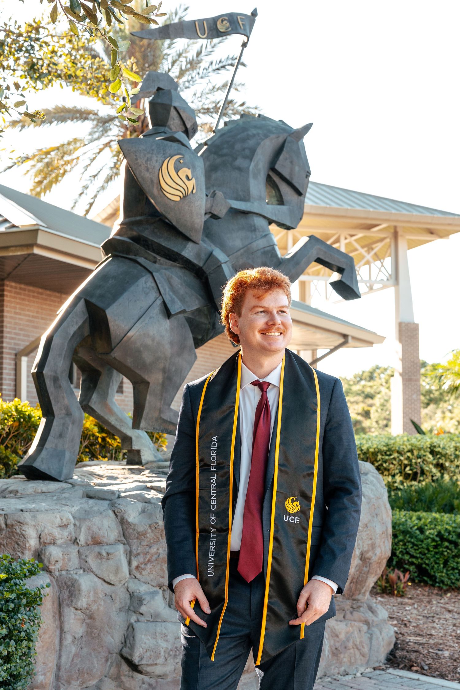 Graduate in cap and gown poses in front of UCF Knight statue during commencement at University of Central Florida.