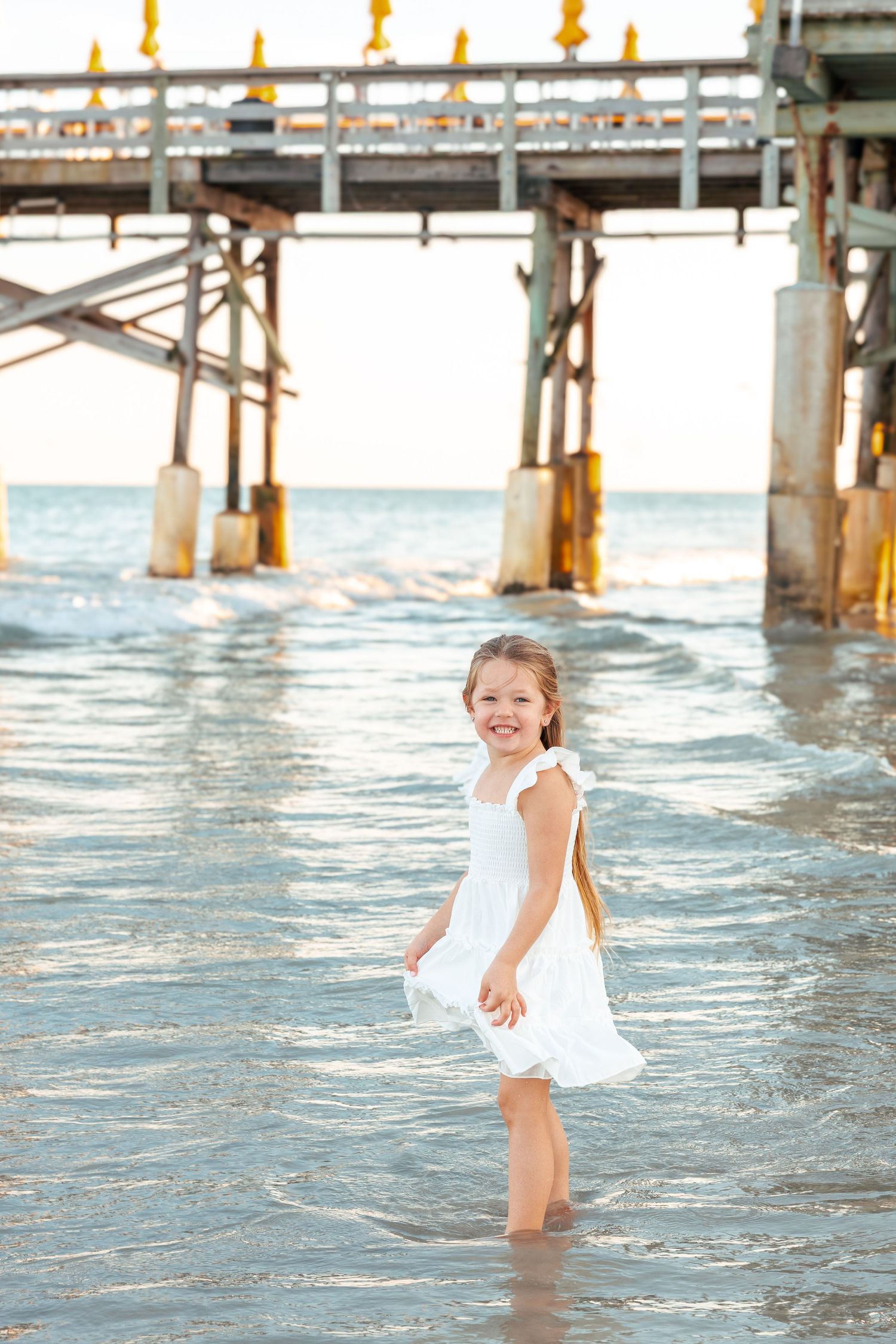 A child in a white dress plays and splashes in the ocean water under a wooden pier on a sunny day.