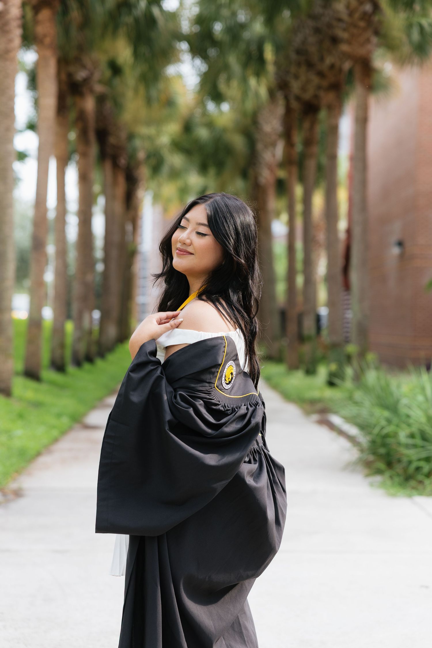 A young woman in a black graduation gown smiles over her shoulder on a palm tree-lined pathway.