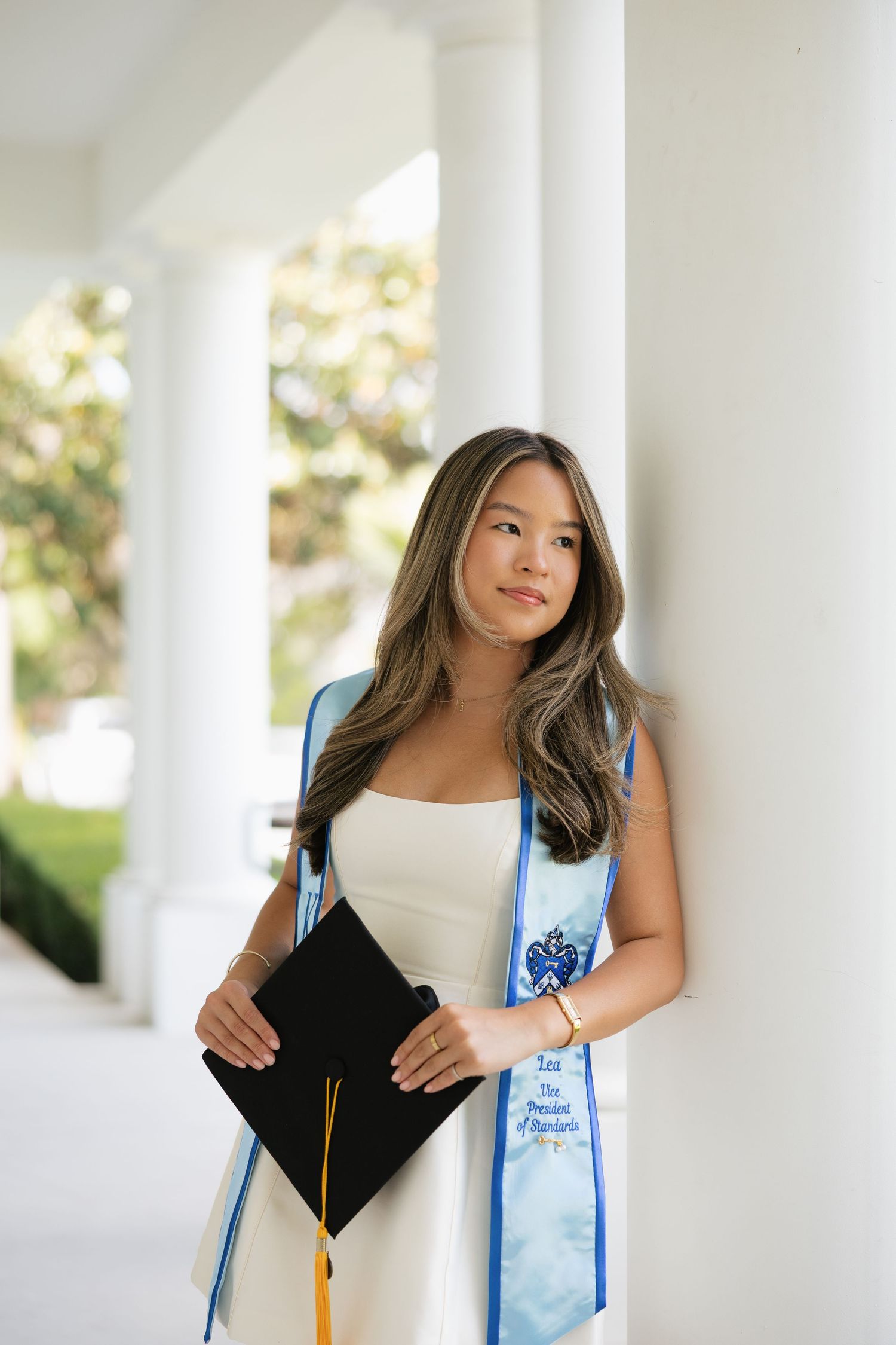 Young woman in white dress holding graduation cap, wearing blue academic stole, leaning against white column outdoors.