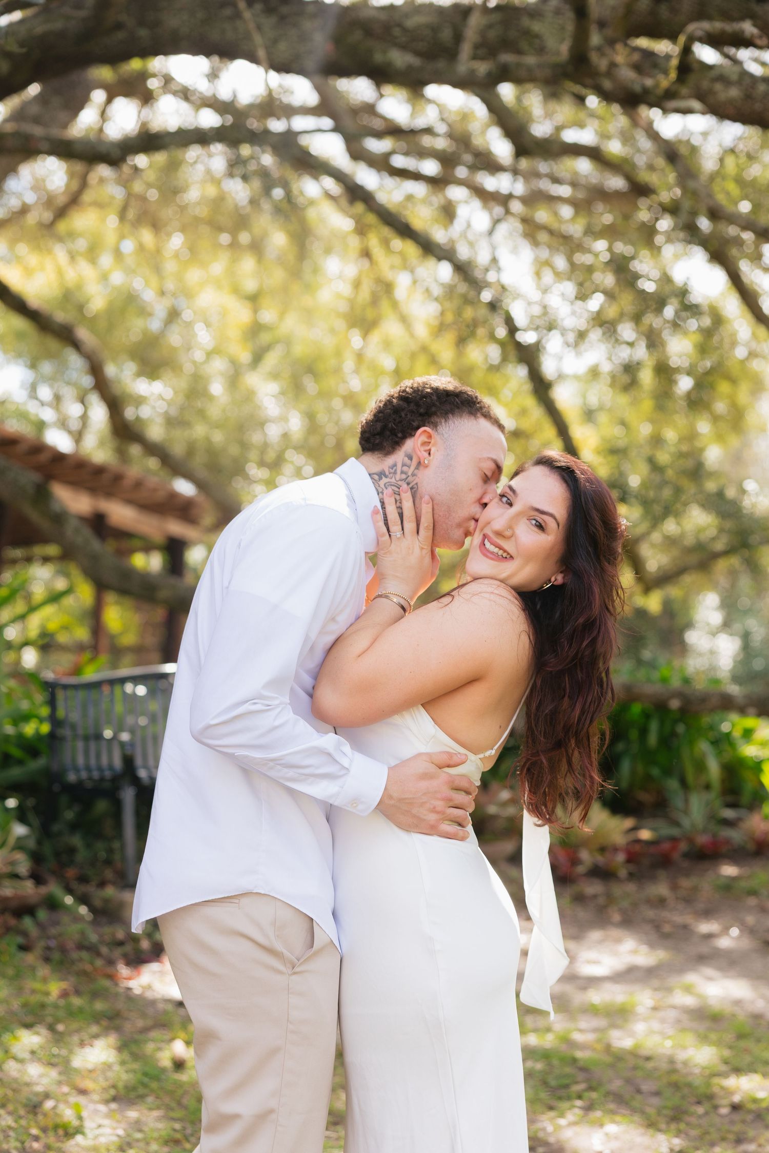 A couple in white attire shares a romantic kiss outdoors under a large tree in a lush garden setting.