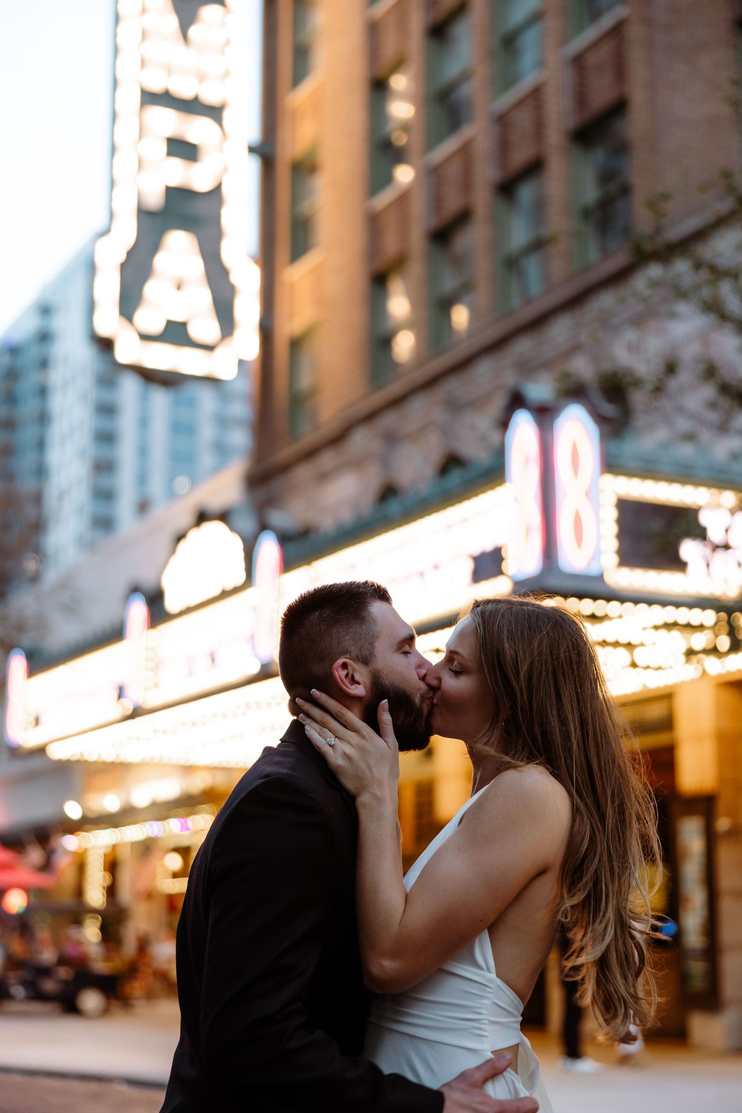 A couple shares a romantic kiss on a city street with illuminated storefronts and buildings in the background.