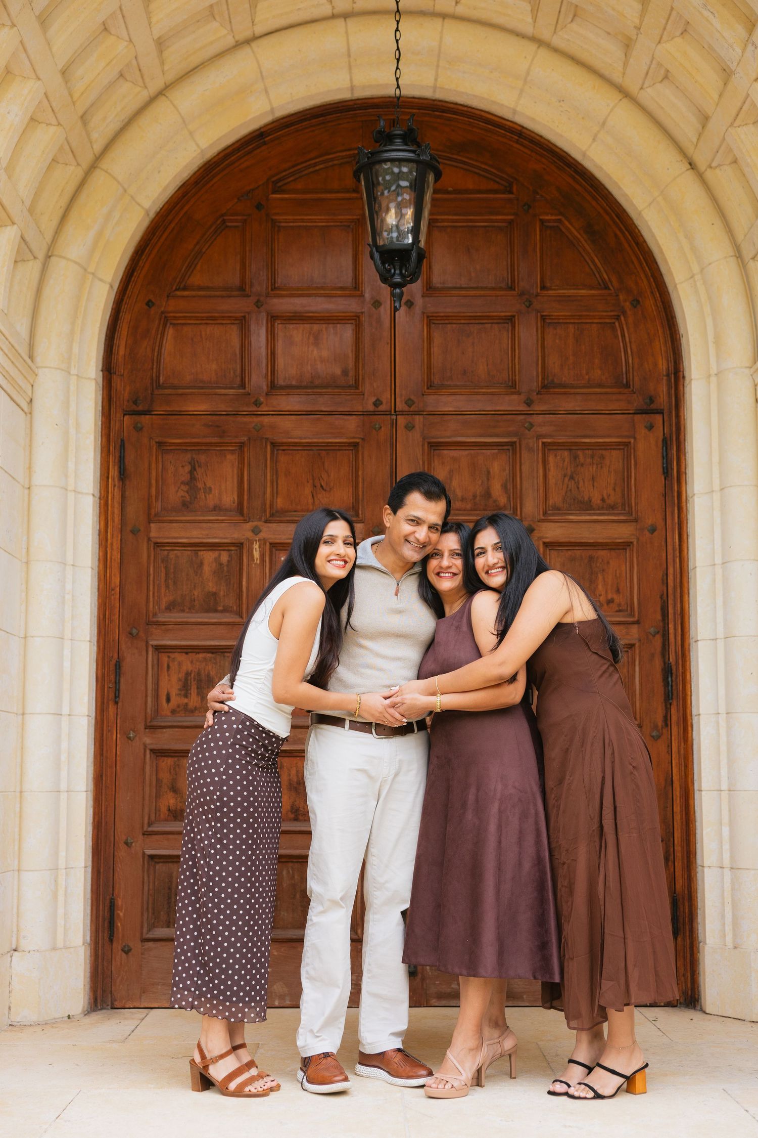 A group stands together embracing in front of a large wooden door with stone archway and hanging lantern.