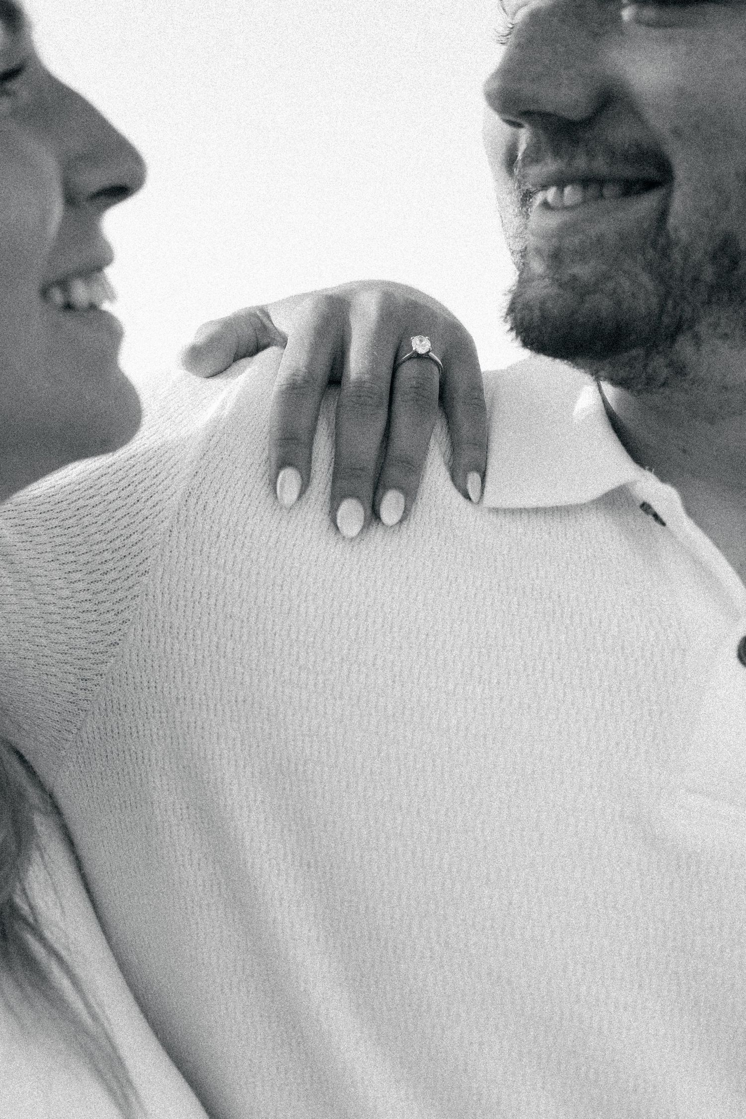 A black and white close-up photo showing a hand with an engagement ring resting on someone's shoulder.